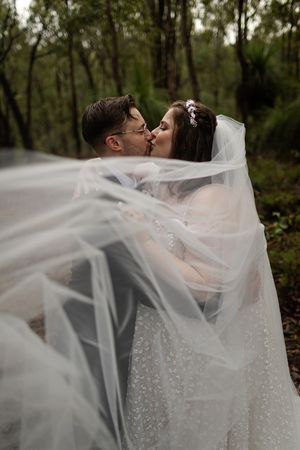 Flowing white veil creates romantic silhouette of newlyweds kissing in a wooded setting.