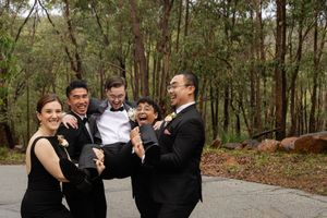 groomsmen in formal black attire laugh together lifting the groom off his feet