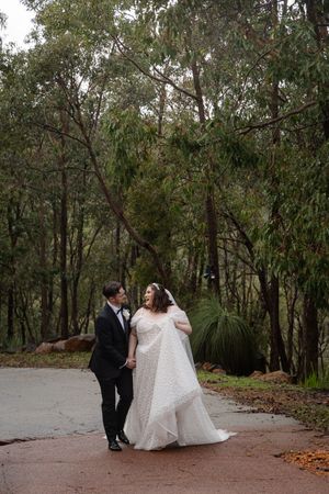 A couple in formal wedding attire walk along a winding path through eucalyptus trees on their special day.