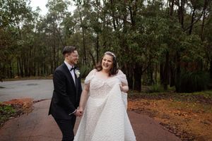 A romantic couple in formal attire walking together on a tree-lined path covered in autumn leaves at a wedding venue.