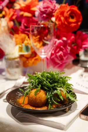 Plated fine dining dish with arugula salad and crispy breaded item on dark plates with vibrant floral decor.