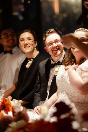 Wedding couple and guests share joyful laughter during reception speeches under warm lighting.