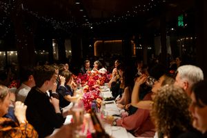 Wedding celebration with guests dining at long tables decorated with red floral centerpieces and twinkling lights.