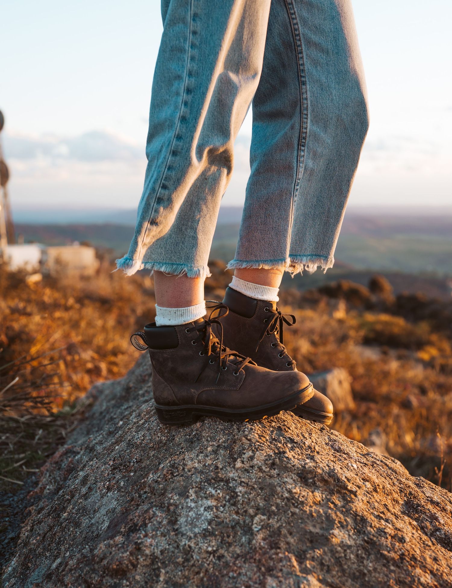 "Person wearing black hiking boots and white socks standing on rocky outcrop at sunset, with distressed denim jeans and golden hour lighting