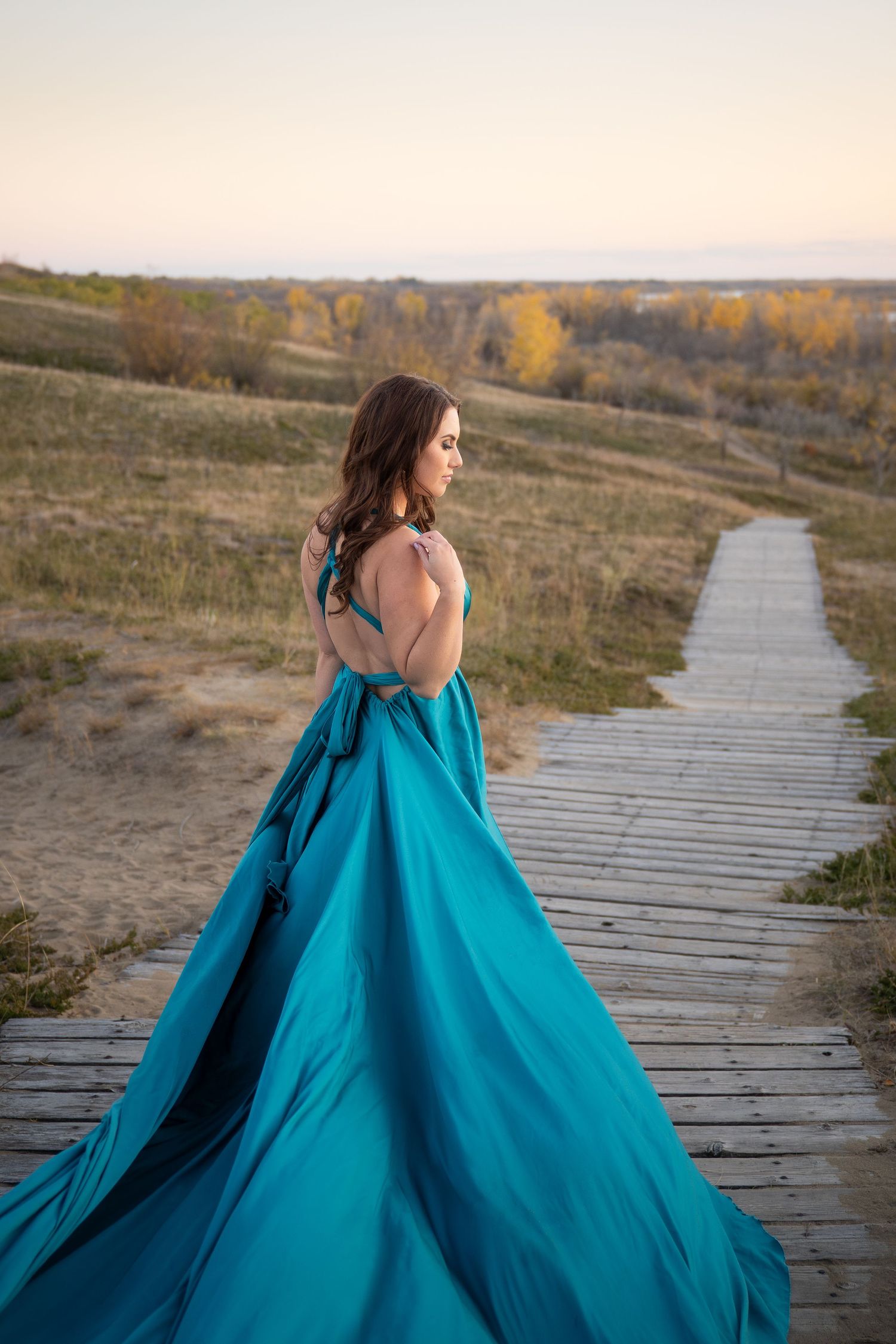 A figure in a flowing turquoise gown walks along a wooden boardwalk through a natural landscape at sunset.