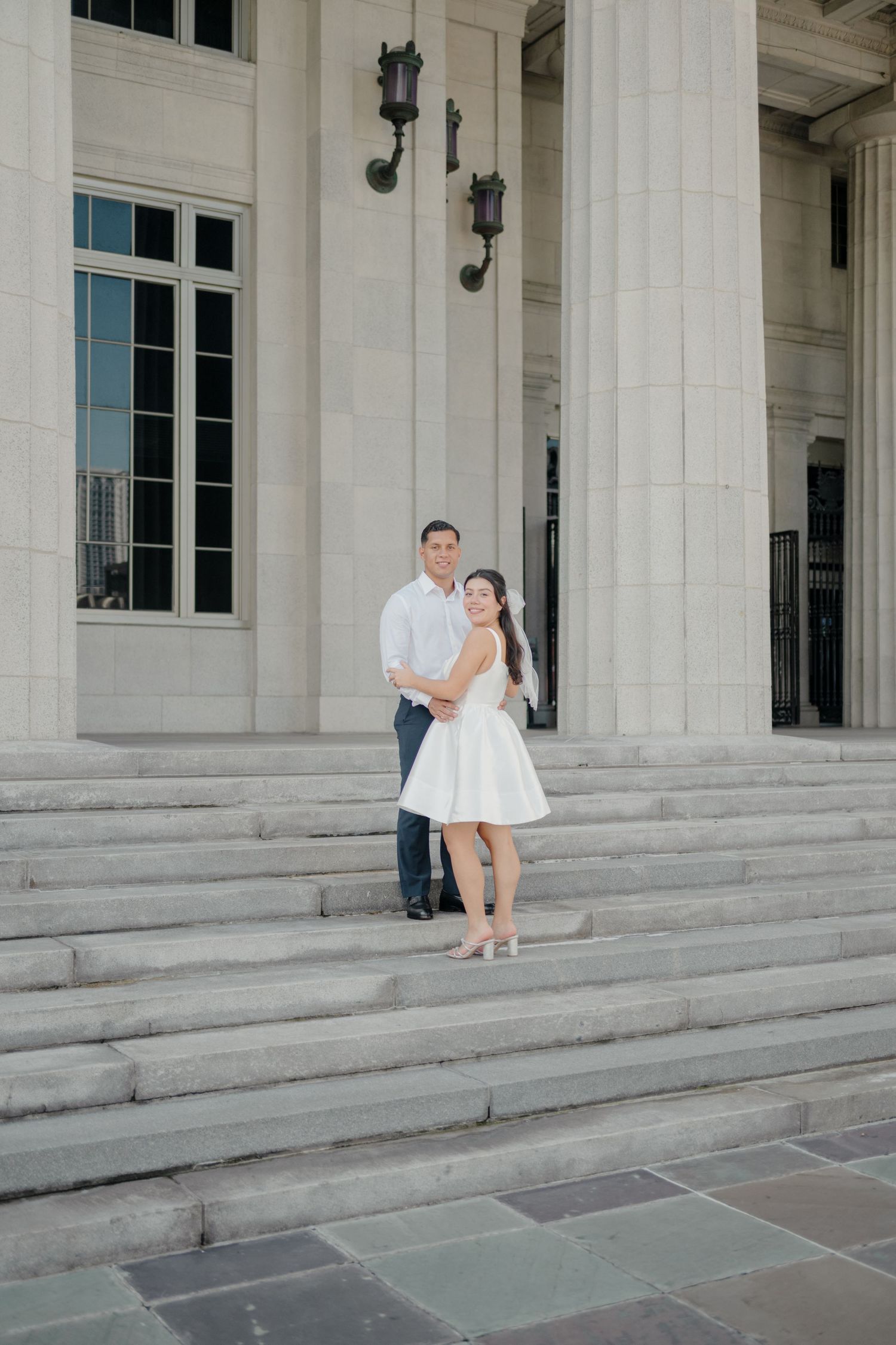 A couple poses playfully on wide stone steps of a neoclassical building with elegant white pillars.