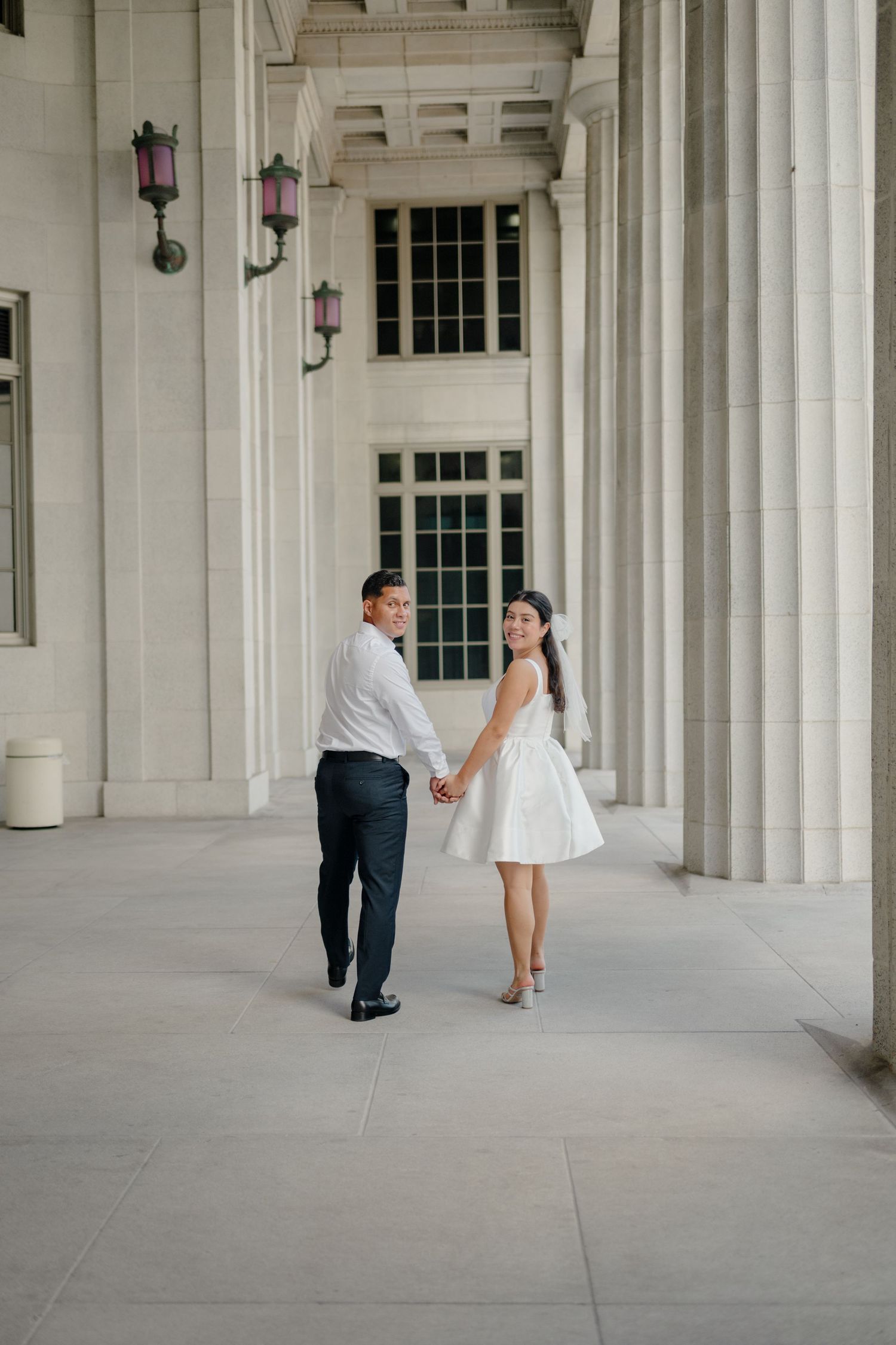 A couple walks through an elegant colonnade with white pillars and archways during a wedding photo session.