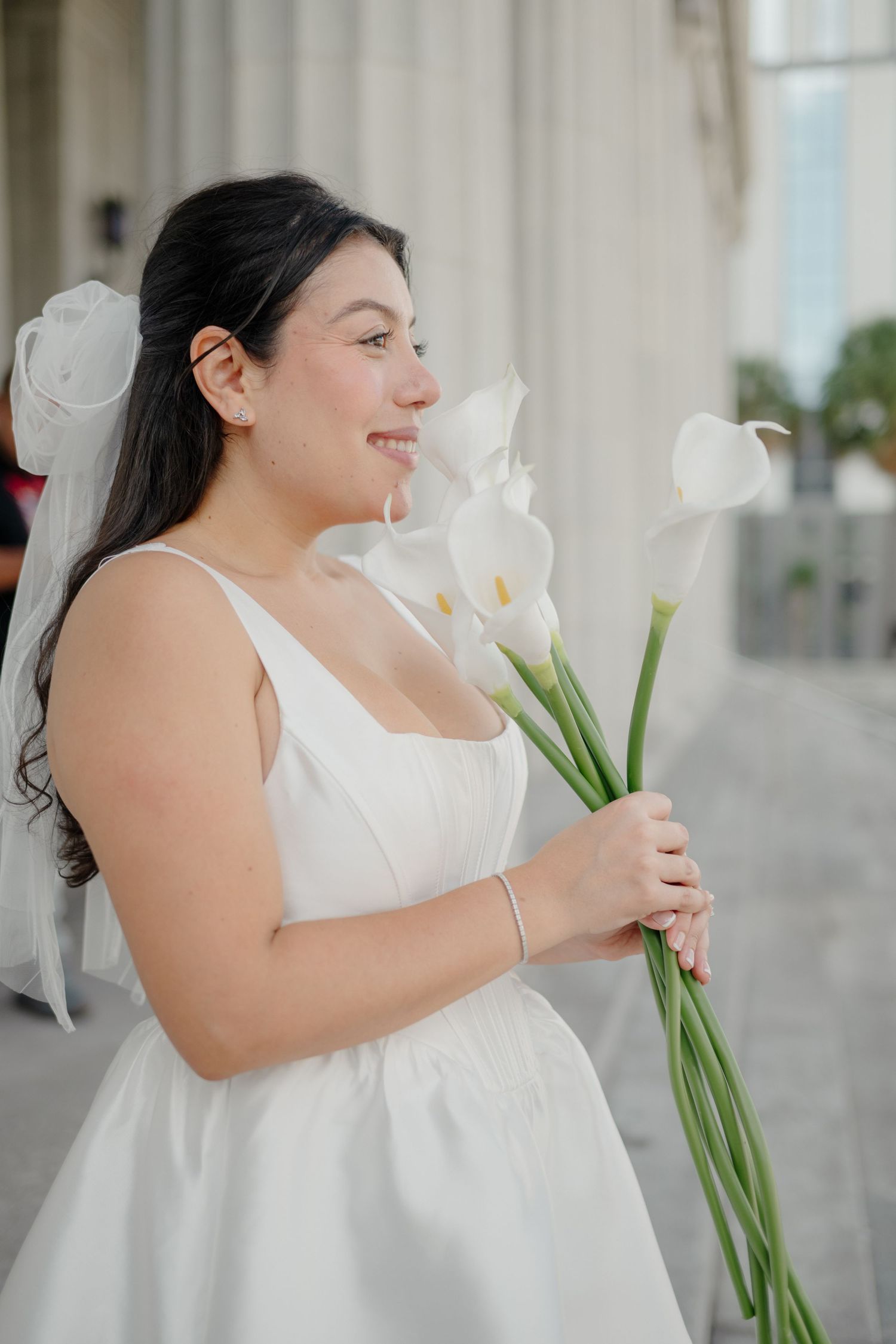 A person in a white wedding dress holds a bouquet of white calla lilies while smiling and looking to the side.