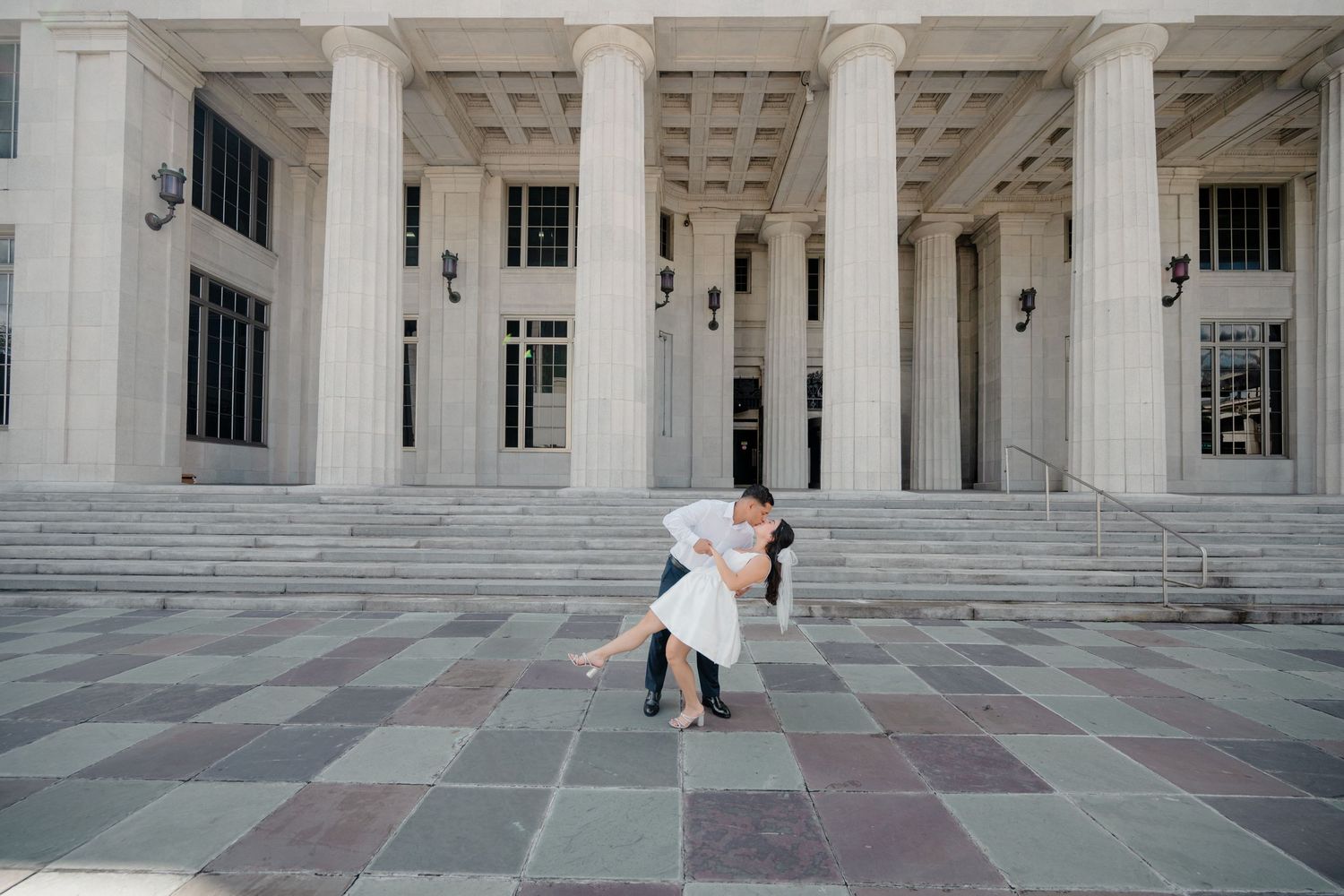 A couple in wedding attire poses on the steps of a grand neoclassical building with white columns.