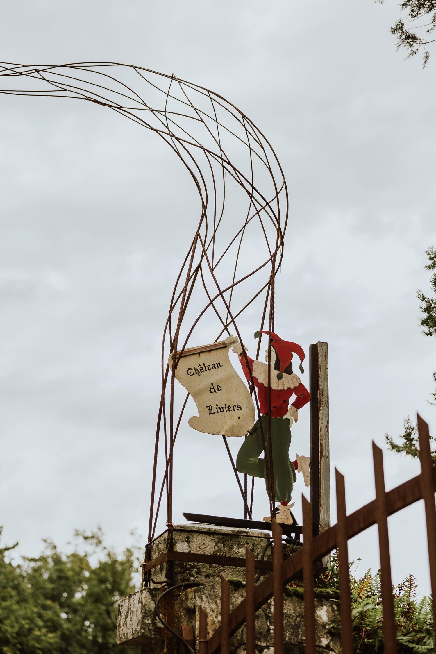 A decorative metal arch with a rustic sign and figure in red perched on wooden posts against an overcast sky.