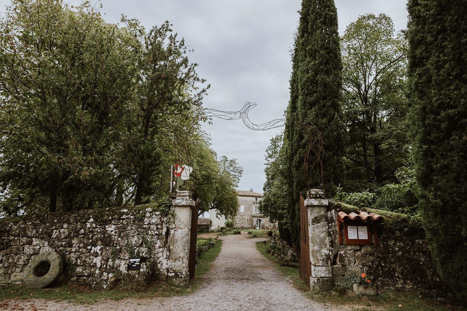 A long gravel pathway lined with tall trees and bushes leads through a peaceful garden setting.