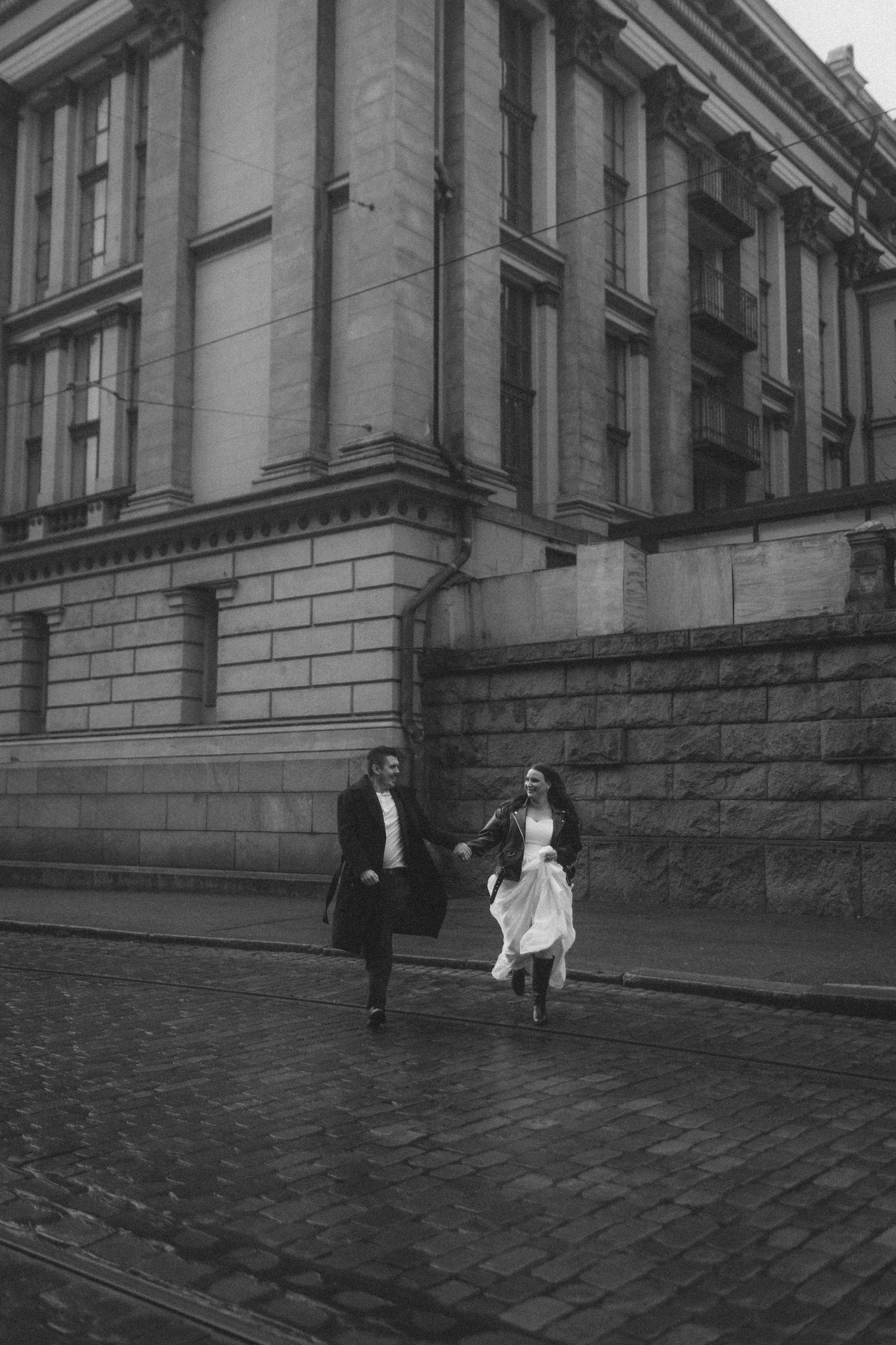 A couple in formal attire dances playfully on cobblestone steps outside a historic building.