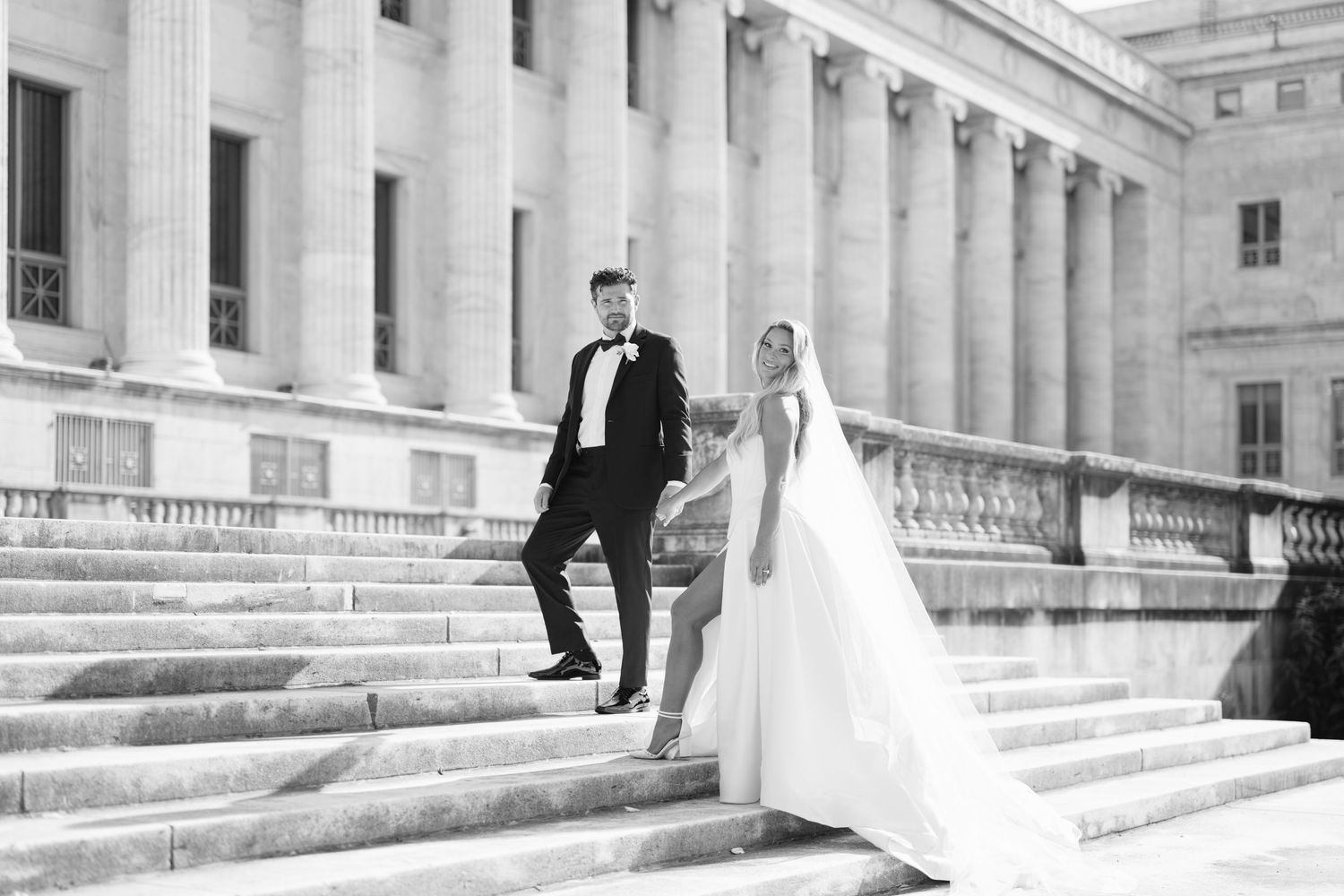 A wedding couple poses on grand stone steps of a classic architectural building in black and white photography.