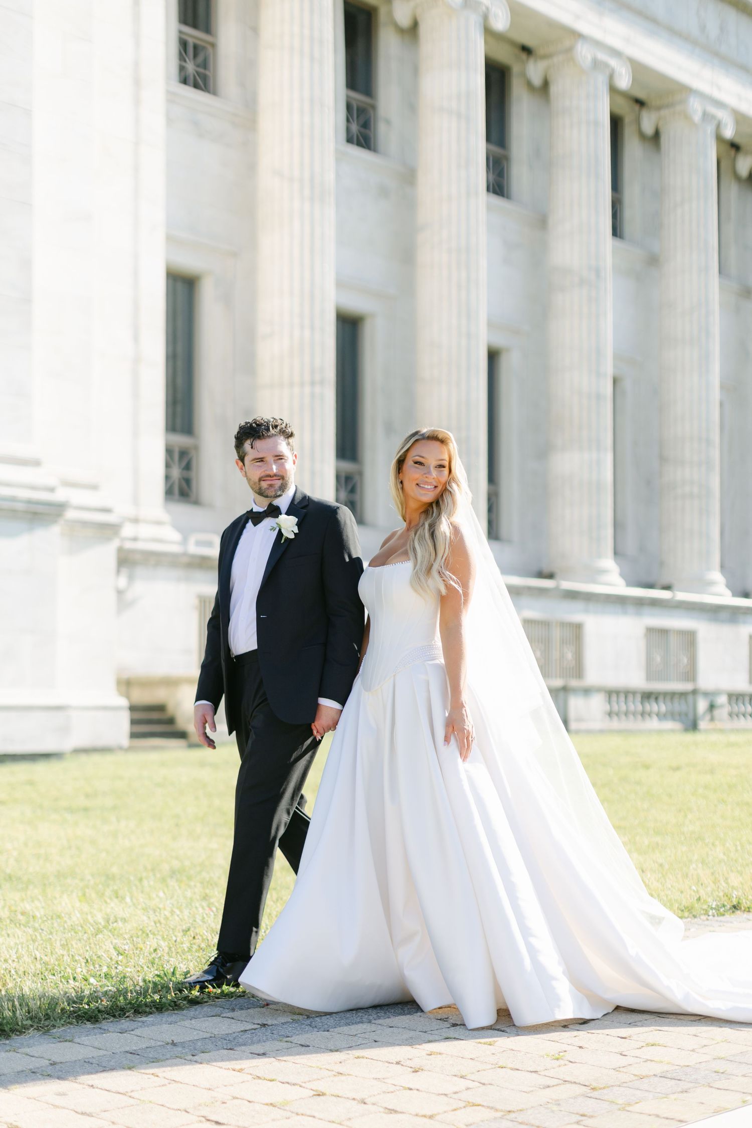 A bride and groom playfully pose on grand white marble steps of a neoclassical building on their wedding day.