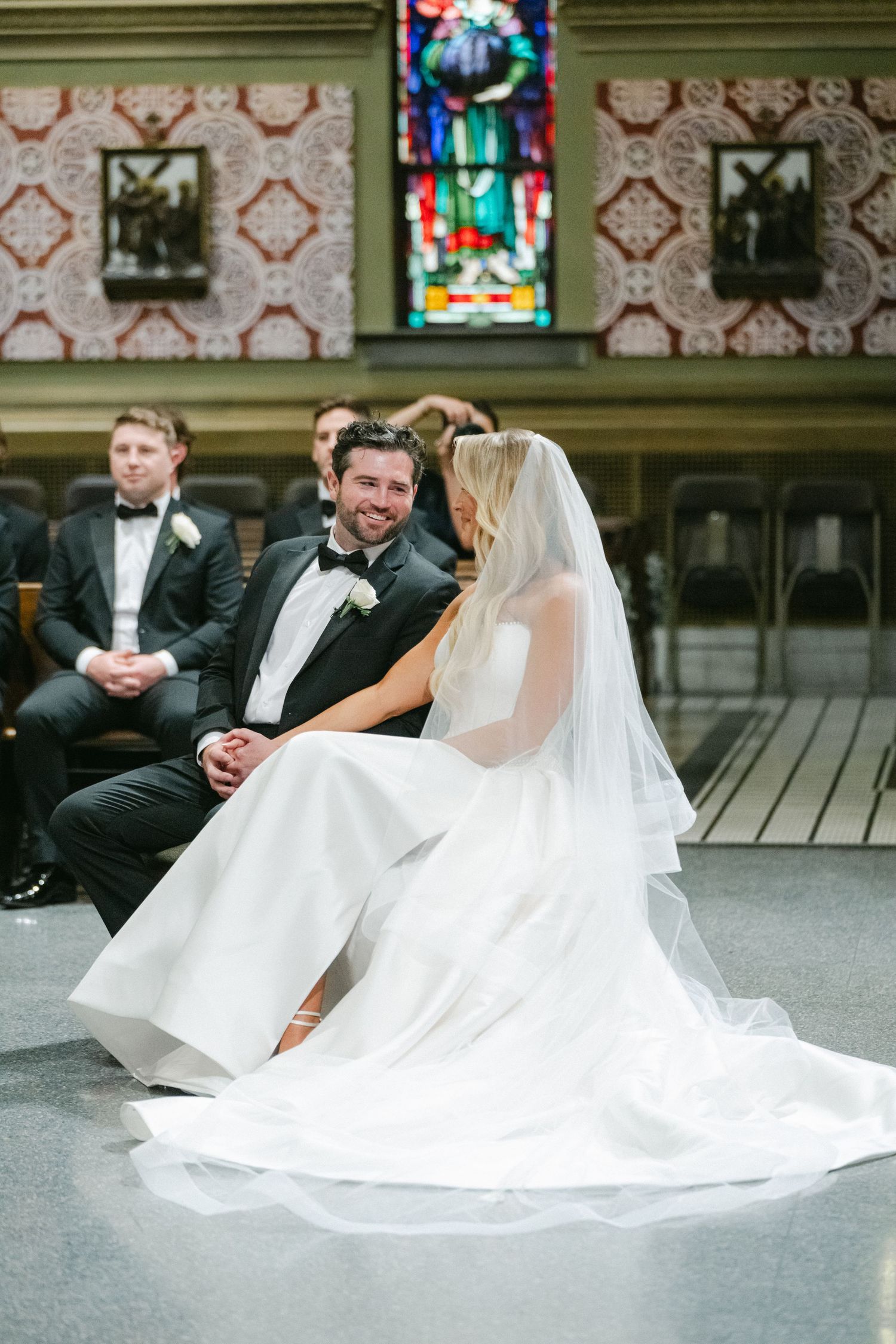 A bride in a flowing white dress sits on wooden steps with her groom in a black tuxedo during their wedding photoshoot.