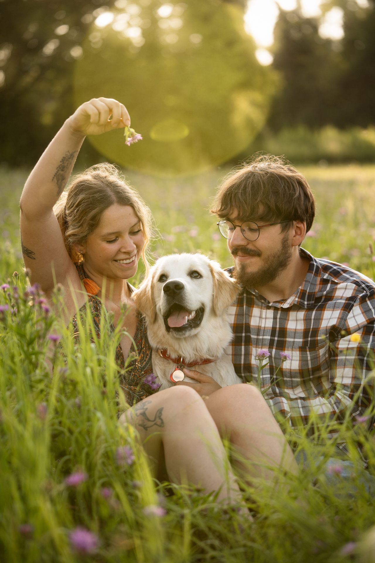 A smiling couple sits together in a wildflower field at sunset with their happy golden retriever dog.