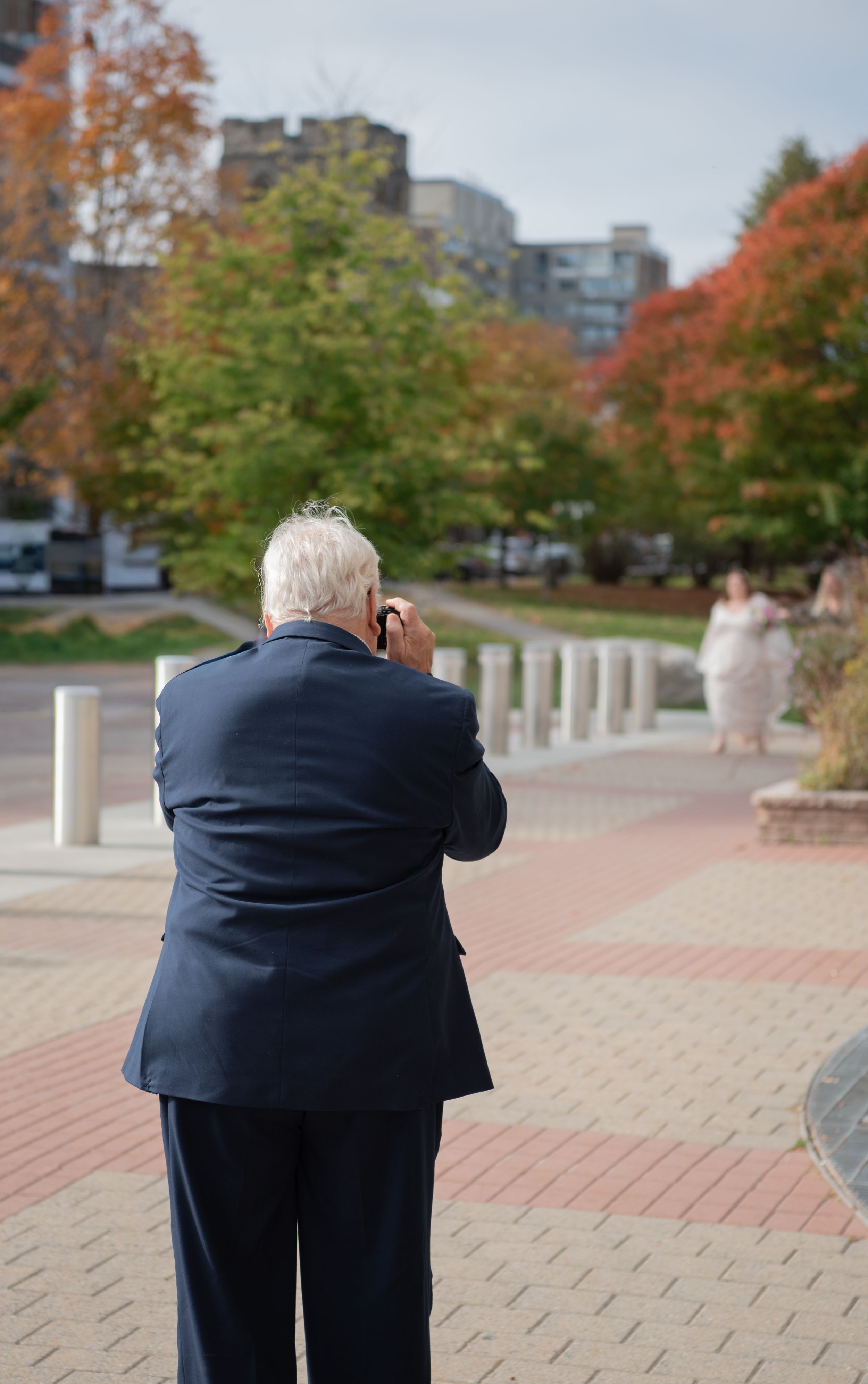 A photographer in a navy suit takes pictures on a brick pathway surrounded by autumn trees and city buildings.