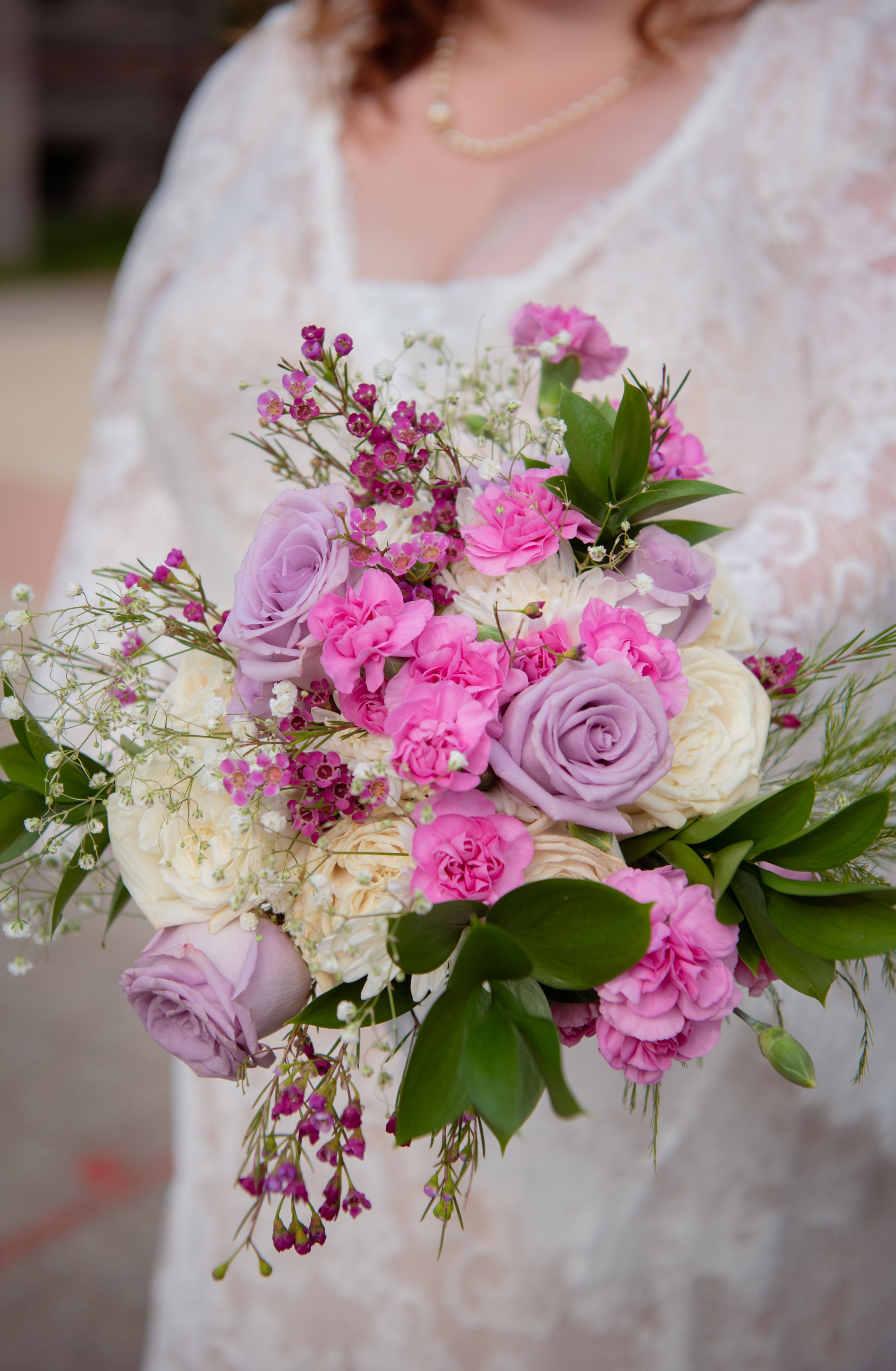 Beautiful bridal bouquet with pink carnations, lavender roses, white hydrangeas and green foliage against lace dress.