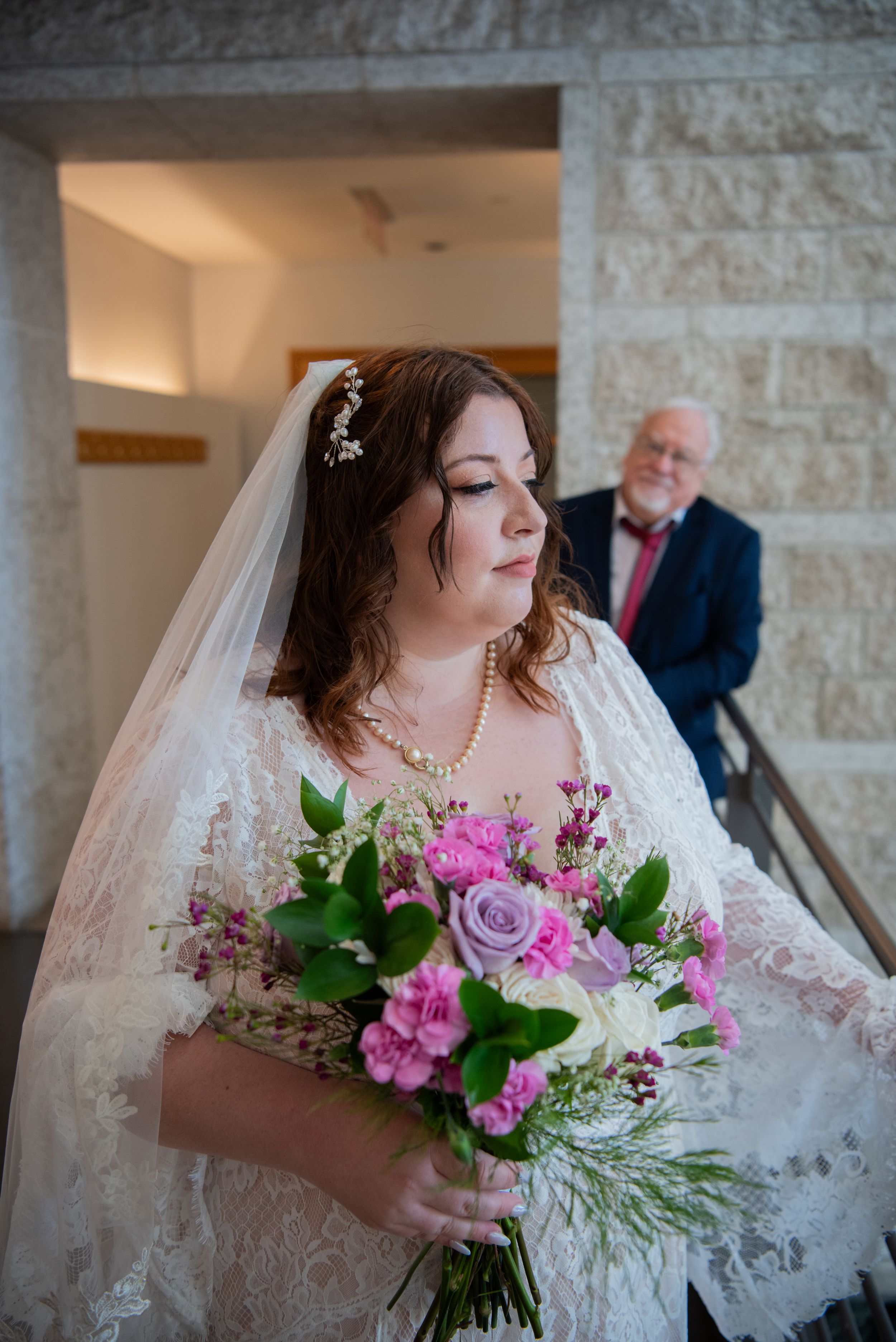 A bride in a white lace gown holds a vibrant pink and purple bouquet against a stone wall interior.