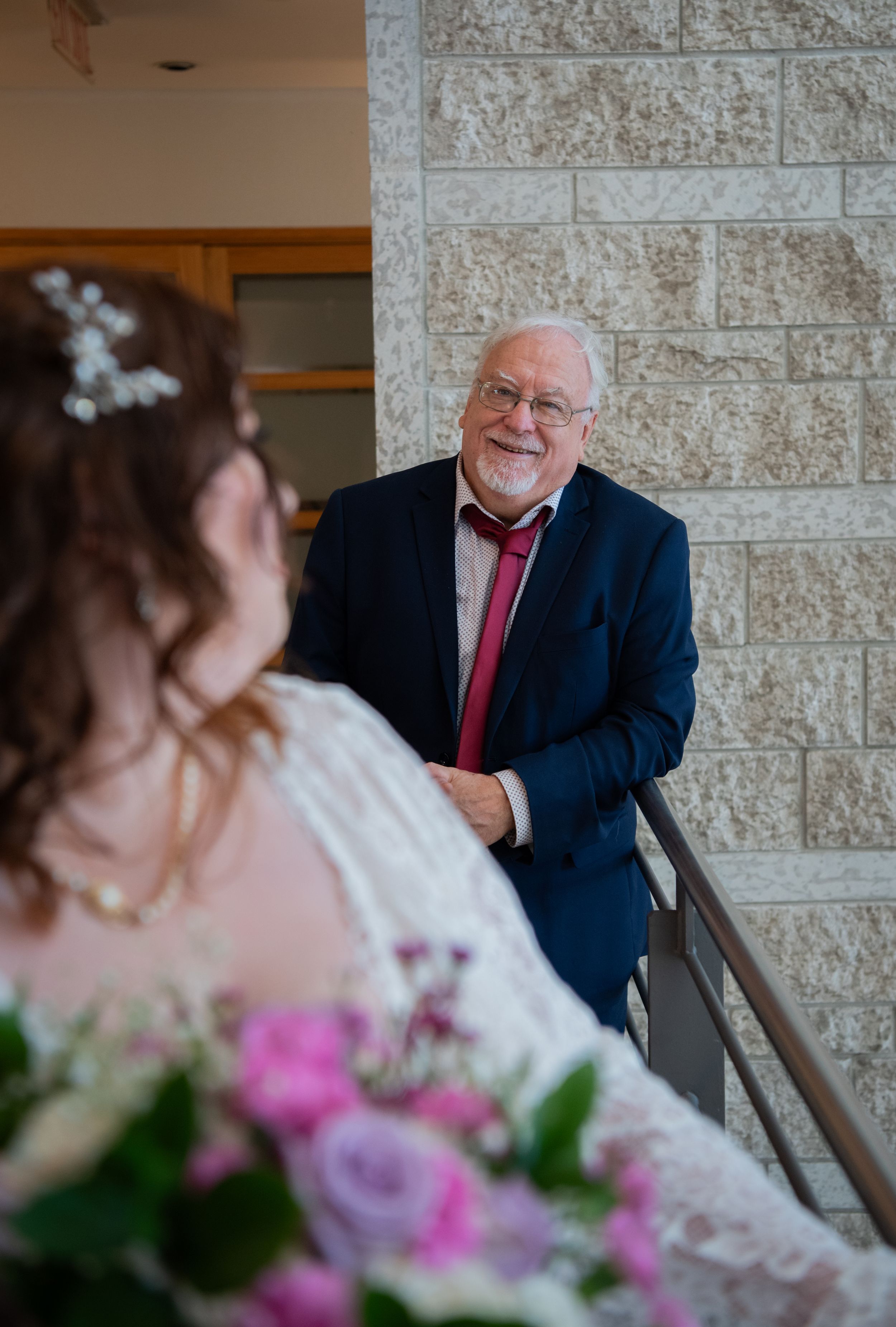 A touching wedding moment captured between someone in a navy suit and red tie and a person in a white lace dress with flowers.