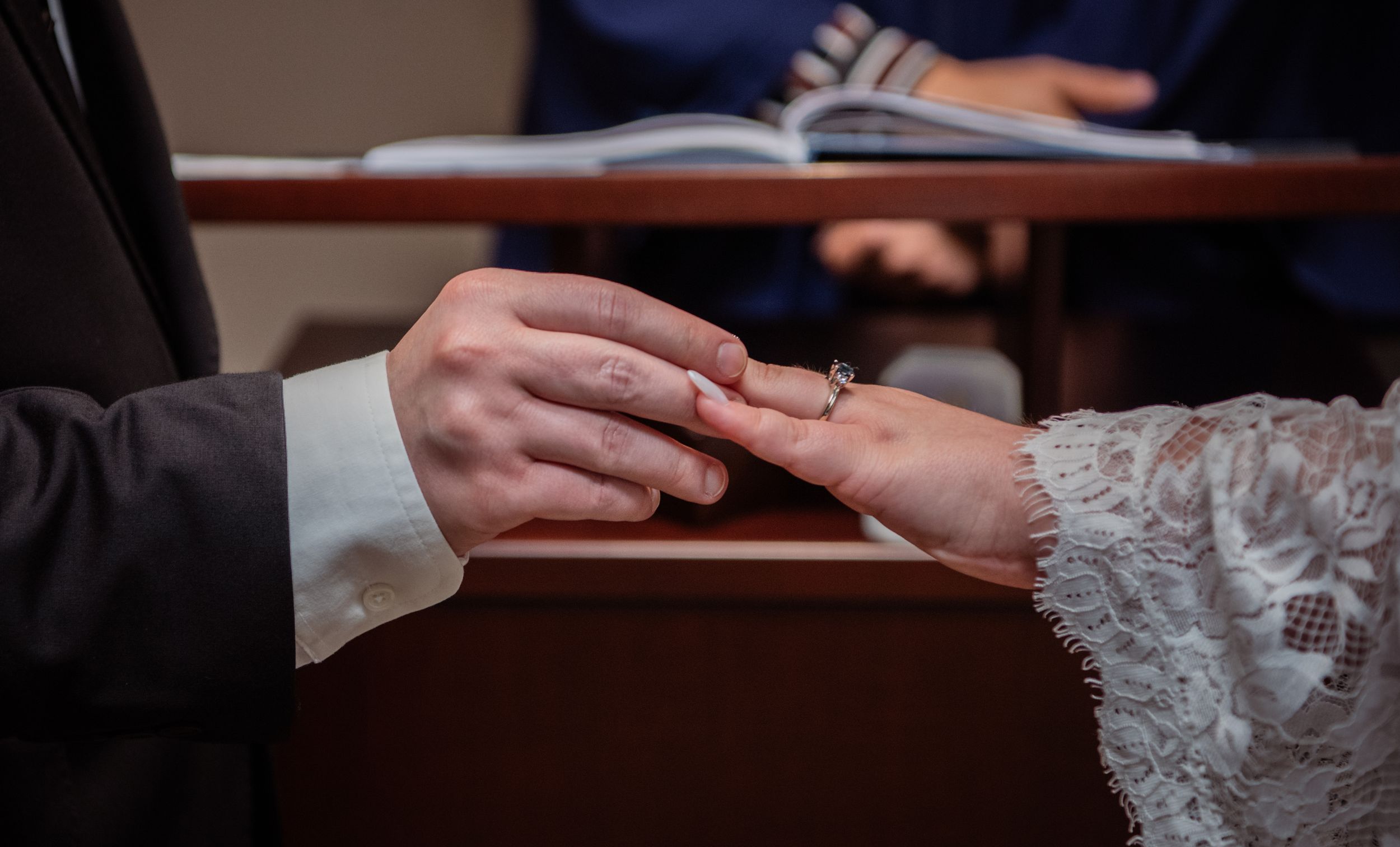 Close-up of hands exchanging wedding rings during marriage ceremony with lace dress detail visible.