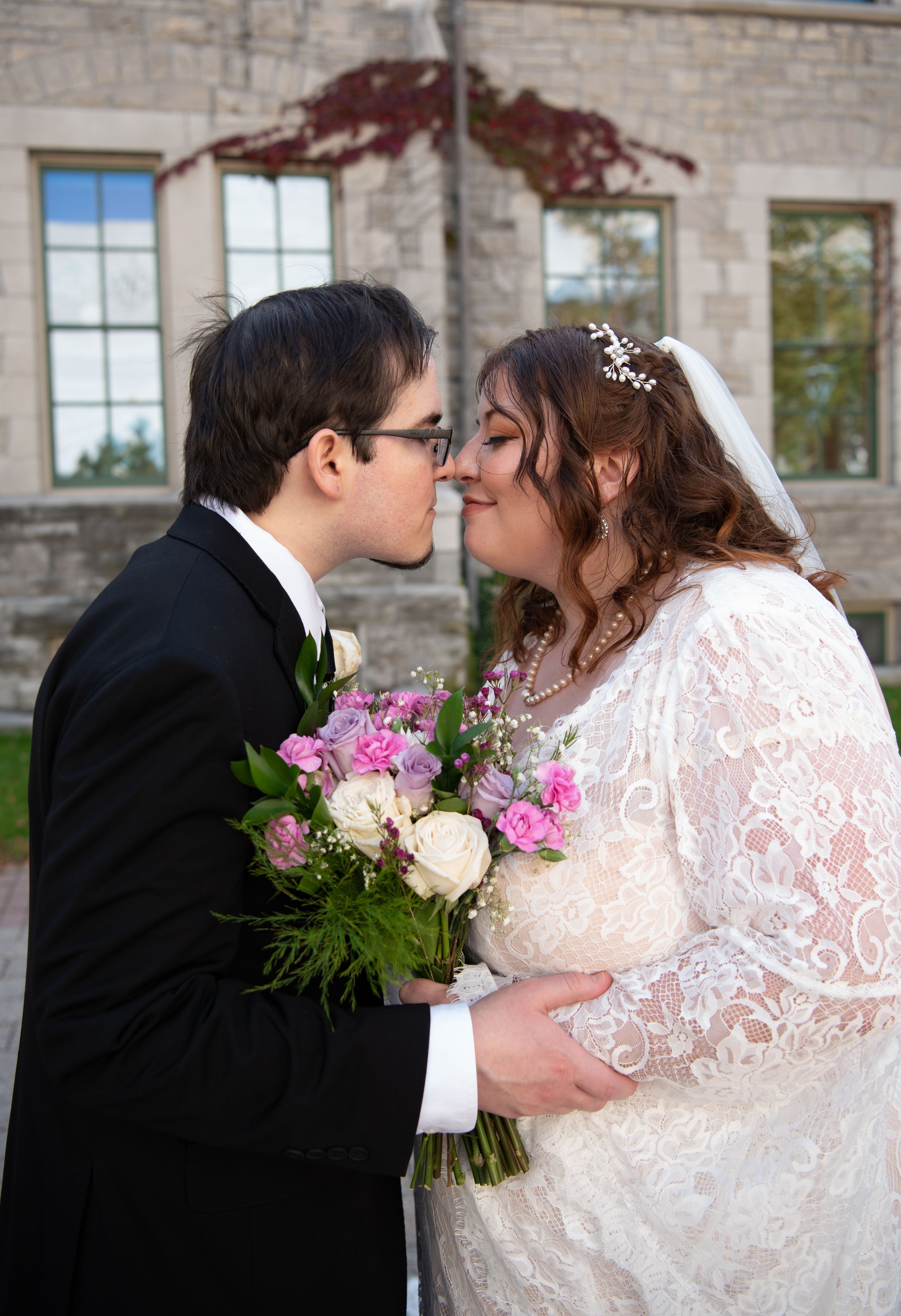 Wedding couple embraces while holding floral bouquet against stone church backdrop.