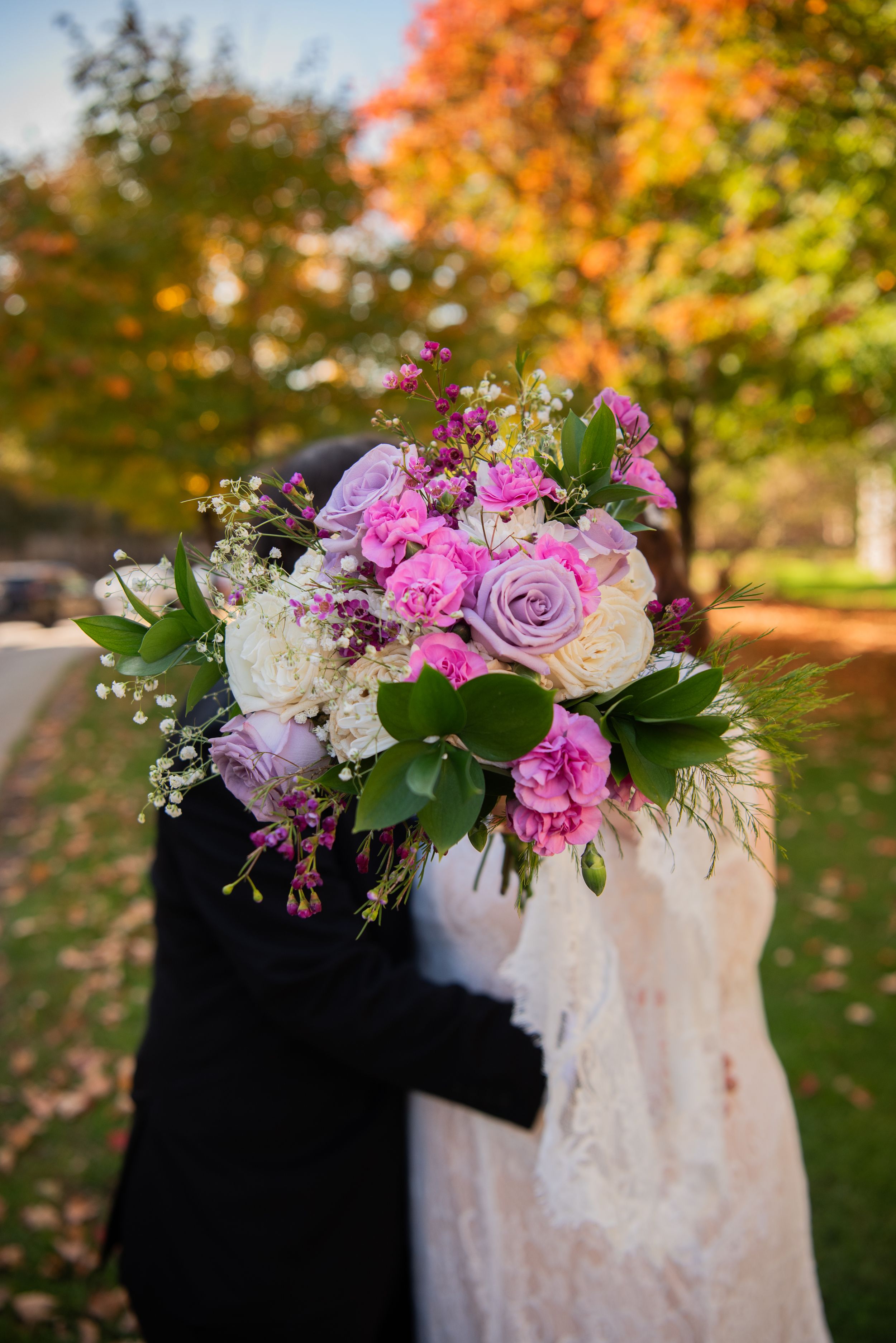 A wedding bouquet with pink roses and purple flowers stands out against autumn foliage in the background.