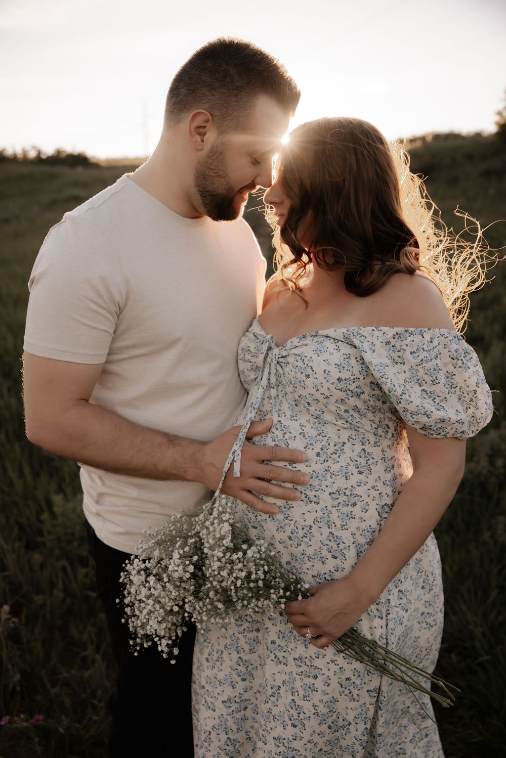 A couple shares a tender moment in a sunlit field, the pregnant woman in a floral dress holding wildflowers at golden hour.