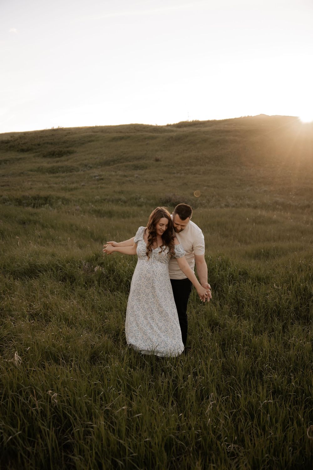 Couple dancing in a sunlit green meadow at golden hour, woman in floral dress, man in white shirt holding hands.