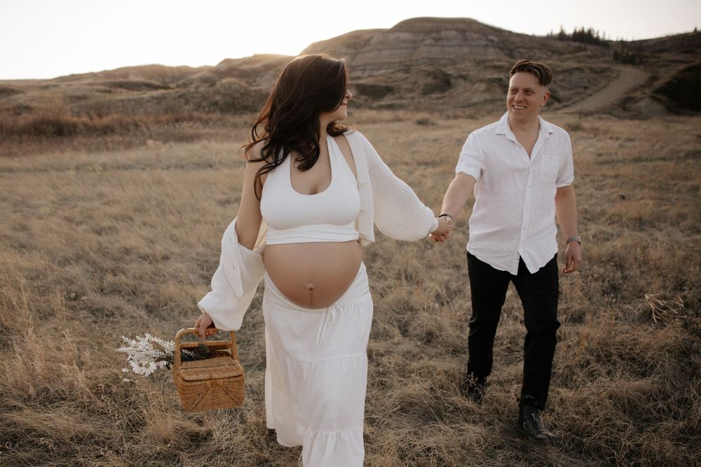 Pregnant woman in white outfit holds picnic basket while walking hand-in-hand with man in field enjoying thier maternity photoshoot