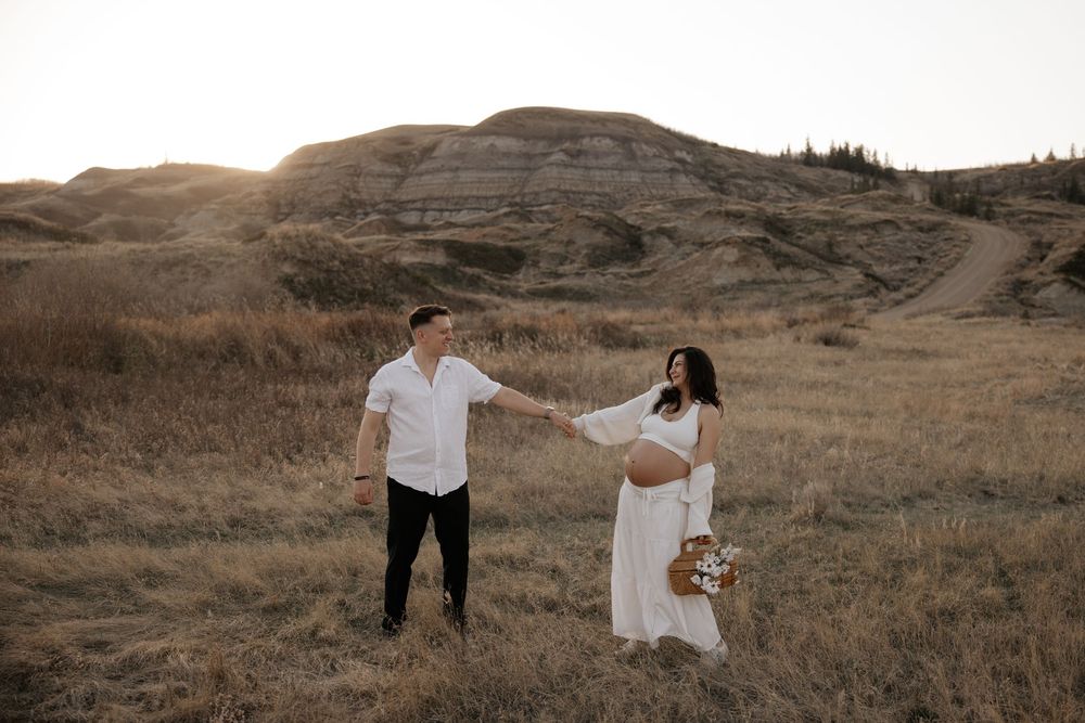 Couple holding hands in golden field in calgary alberta with badlands hills in background during their maternity photography shoot