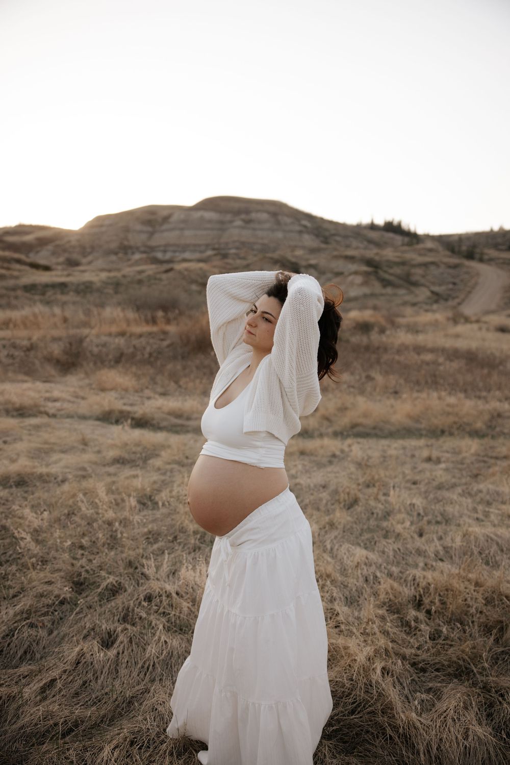 Pregnant woman in white crop top and maxi skirt poses in golden prairie landscape during maternity photoshoot.