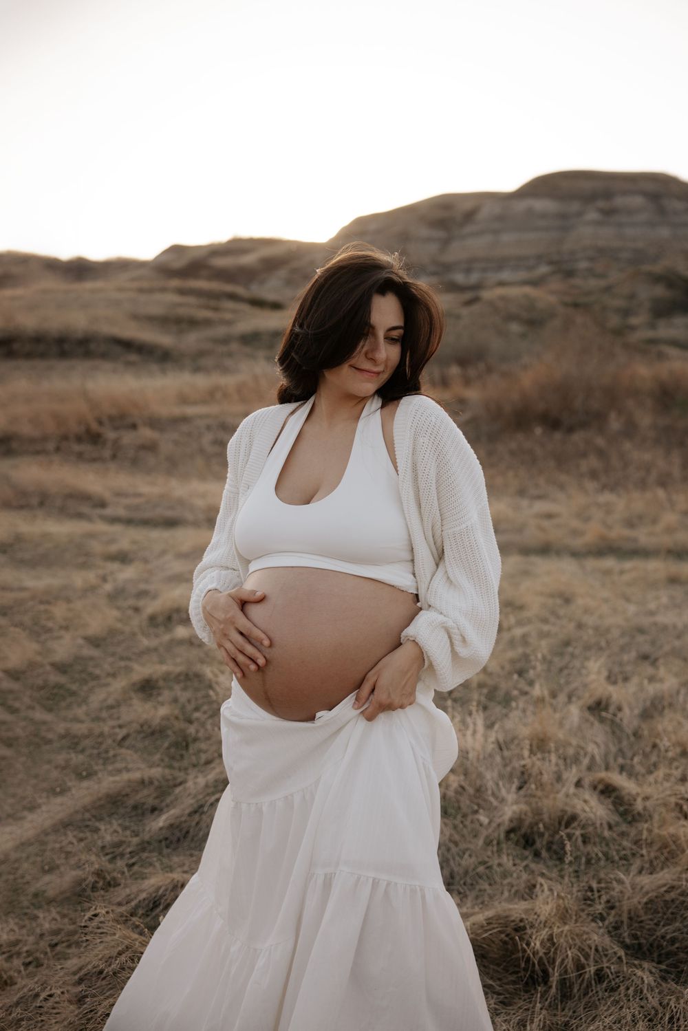 Pregnant woman in white outfit cradling baby bump in golden field at sunset in red deer alberta