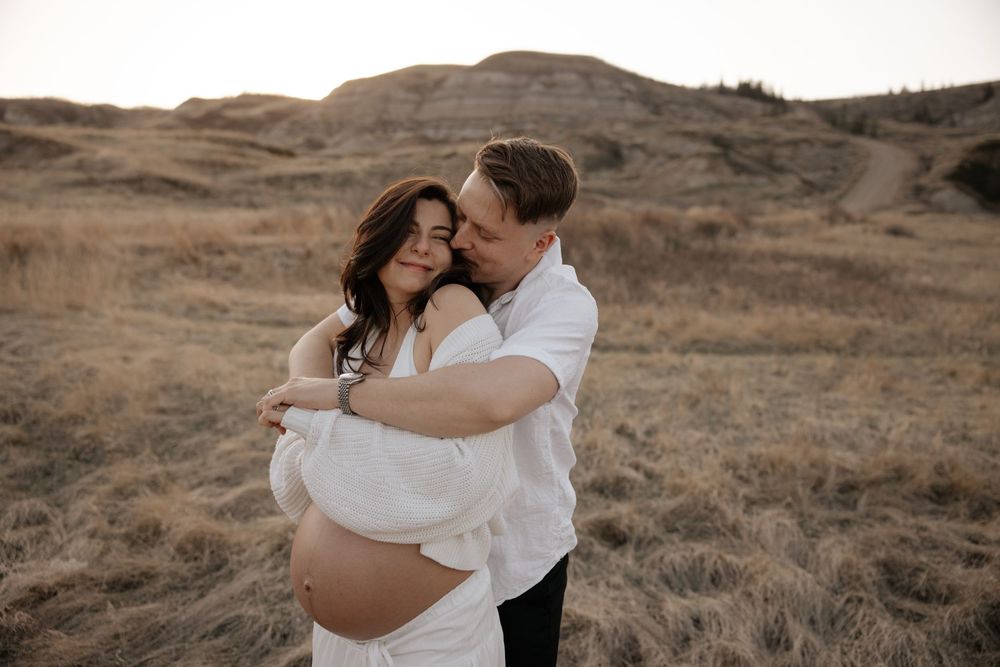 Couple embracing in golden field, pregnant woman in white dress, being embraced by her husband for their maternity photoshoot in calgary