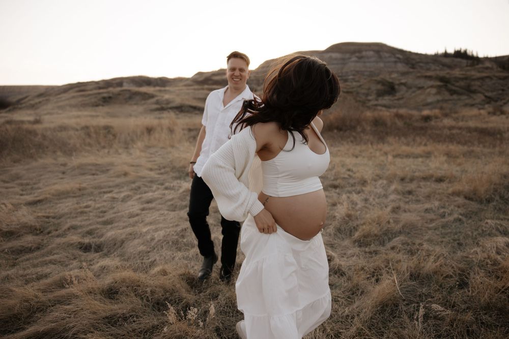 Pregnant woman in white dress holds pregnancy bump runs in a playful way during her maternity shoot in calgary alberta