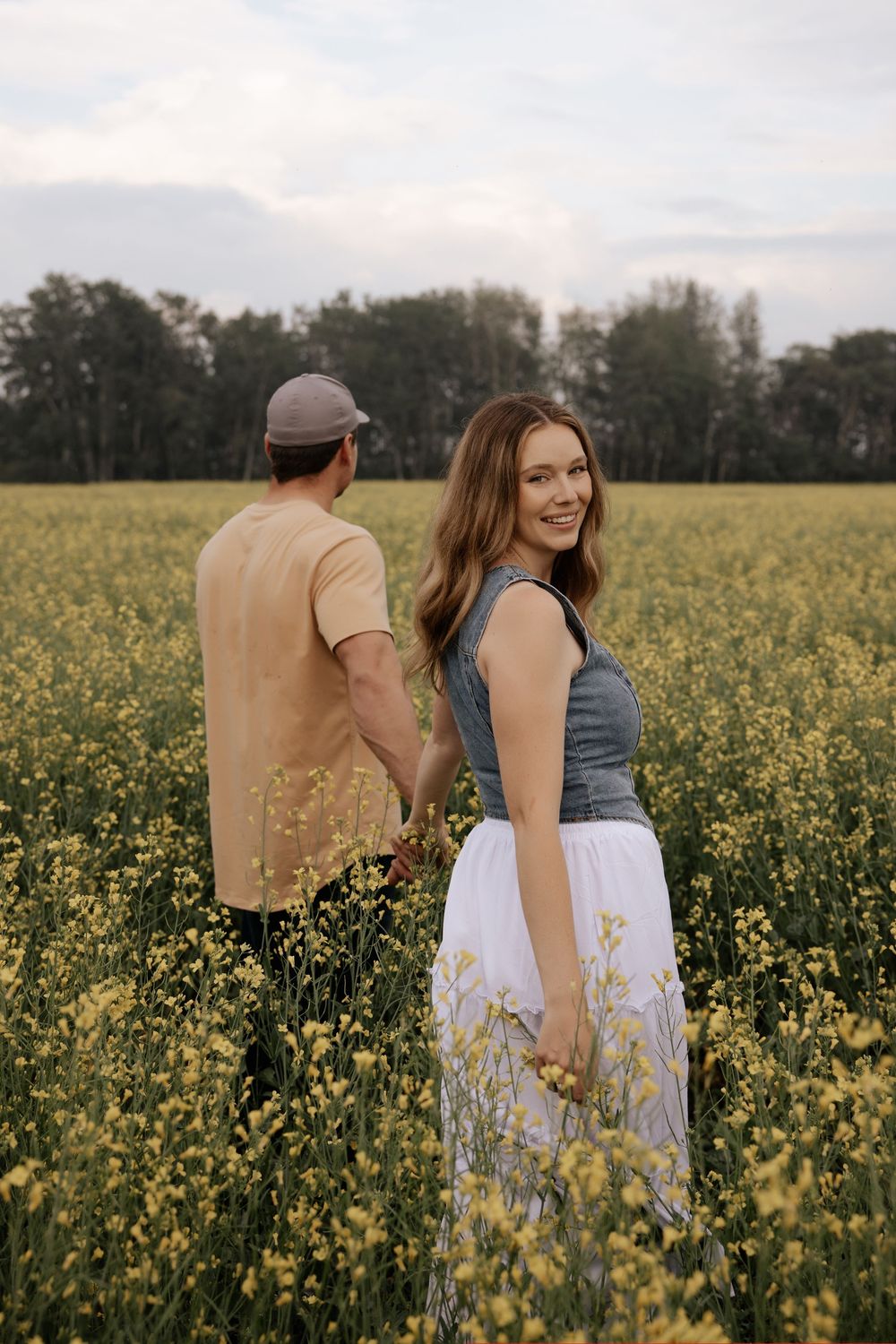 A smiling woman in a gray top and white skirt holds hands with a man in a yellow field of blooming flowers.