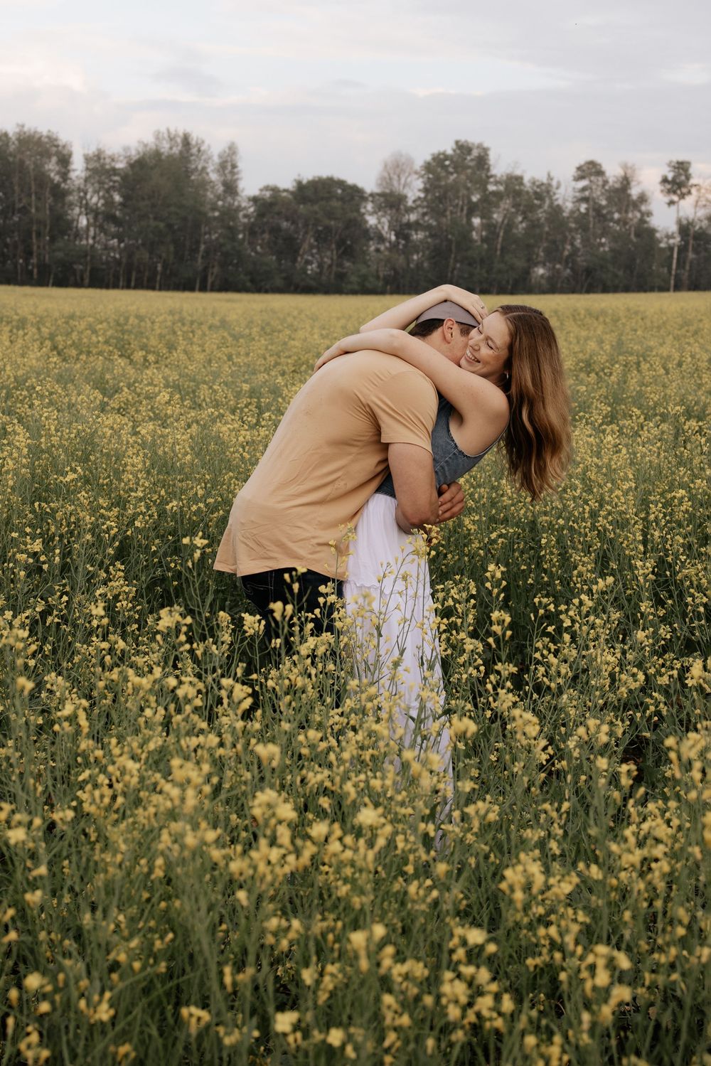 Couple embracing romantically in a vibrant yellow wildflower field with green trees in the background.