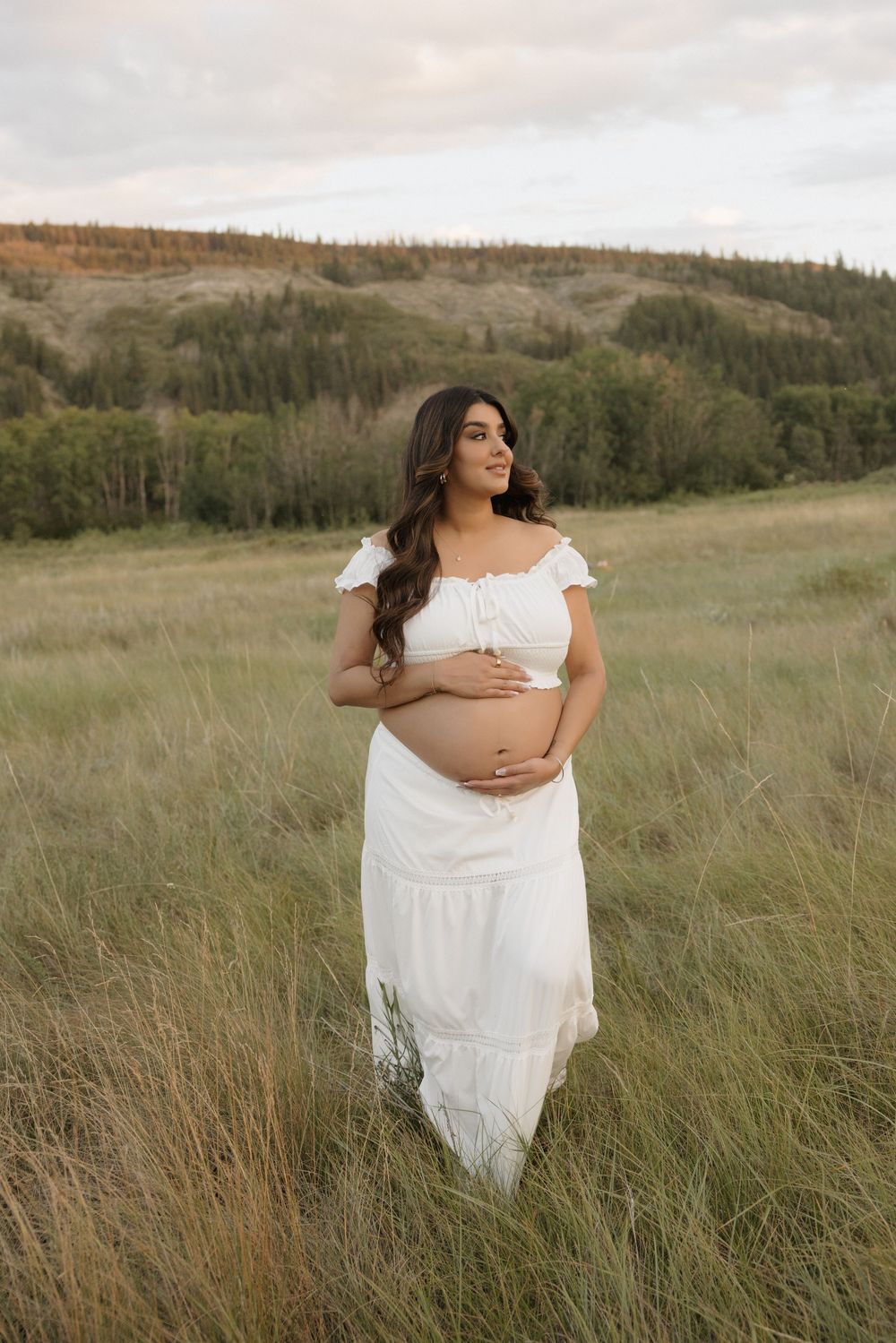 Pregnant woman in white off-shoulder crop top and maxi skirt cradling baby bump during a photoshoot in calgary