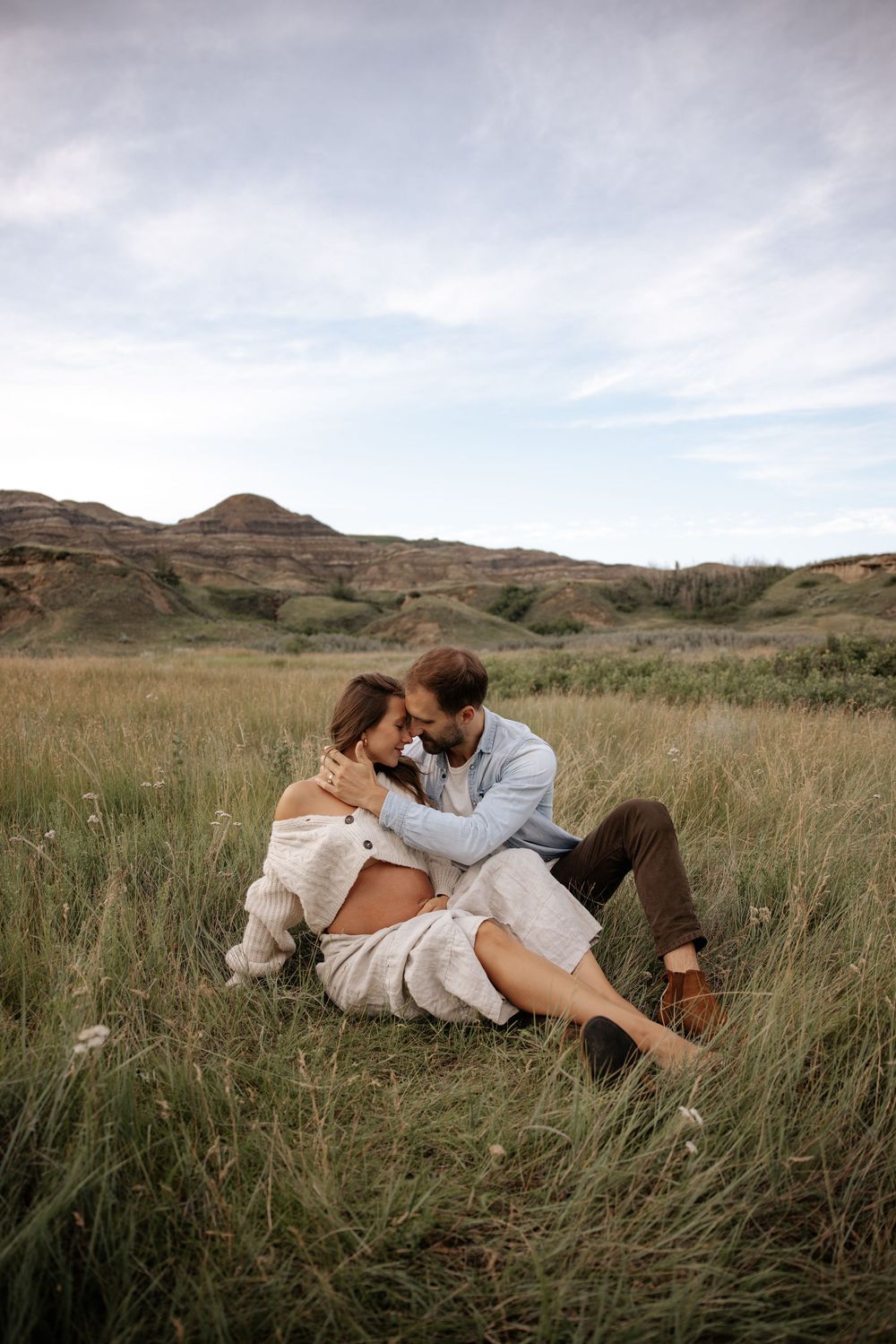 Couple sitting in a grassy field embracing romantically with rolling hills and cloudy sky in the background.