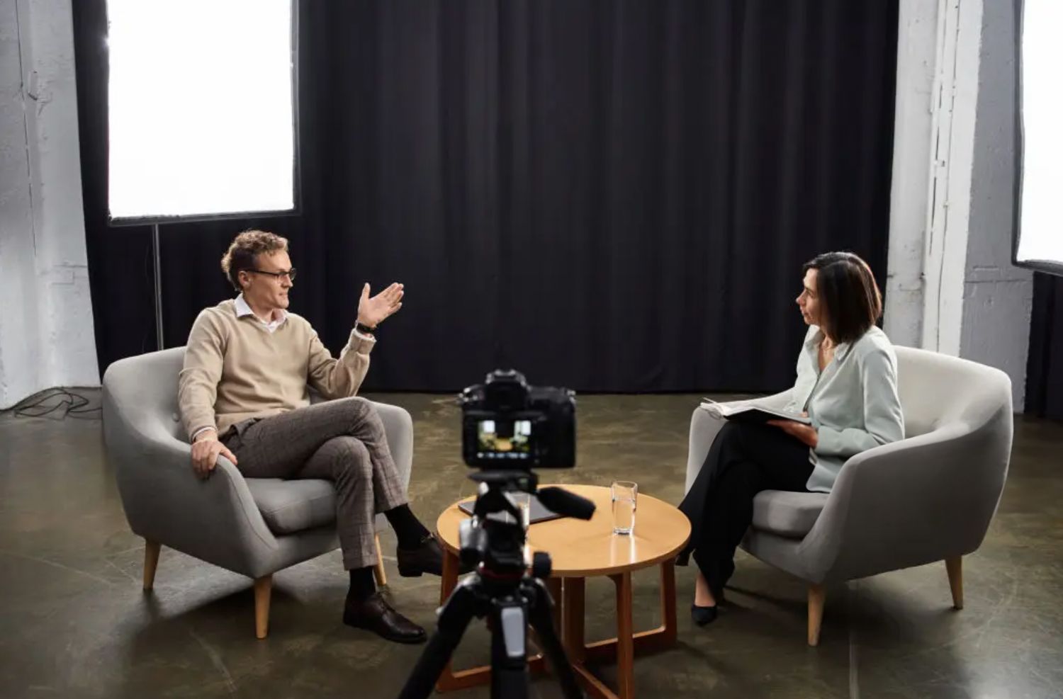 Two people sit in gray armchairs for an interview setup with camera equipment and small table in between them.