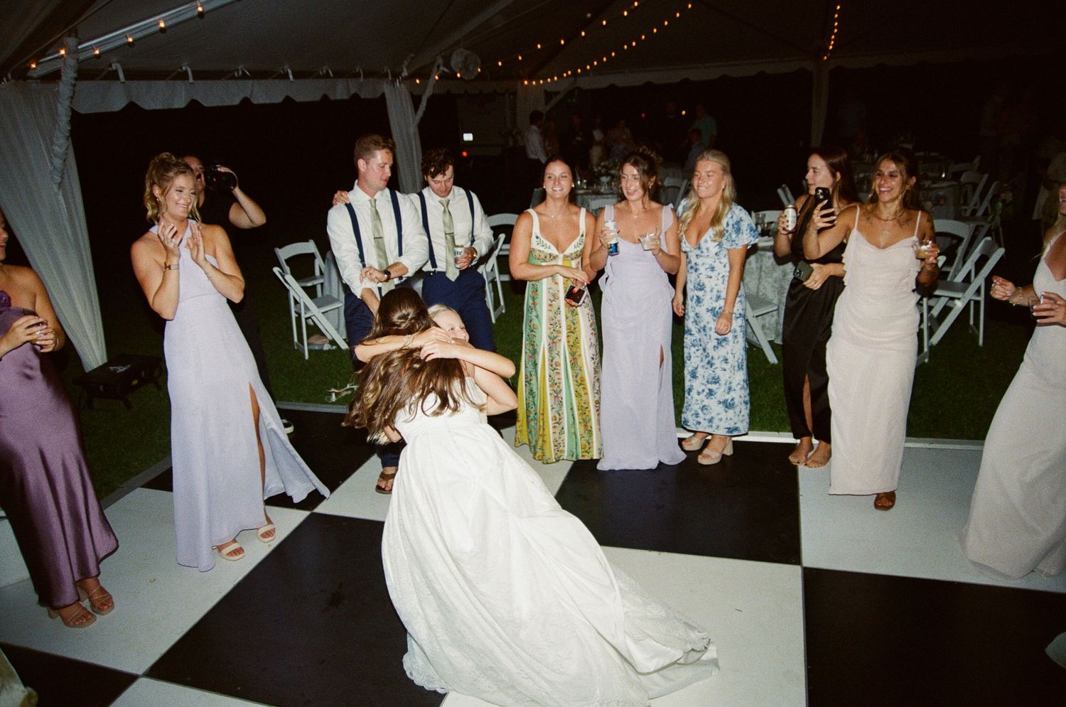 Wedding guests dancing and celebrating on a dark dance floor illuminated by overhead string lights.