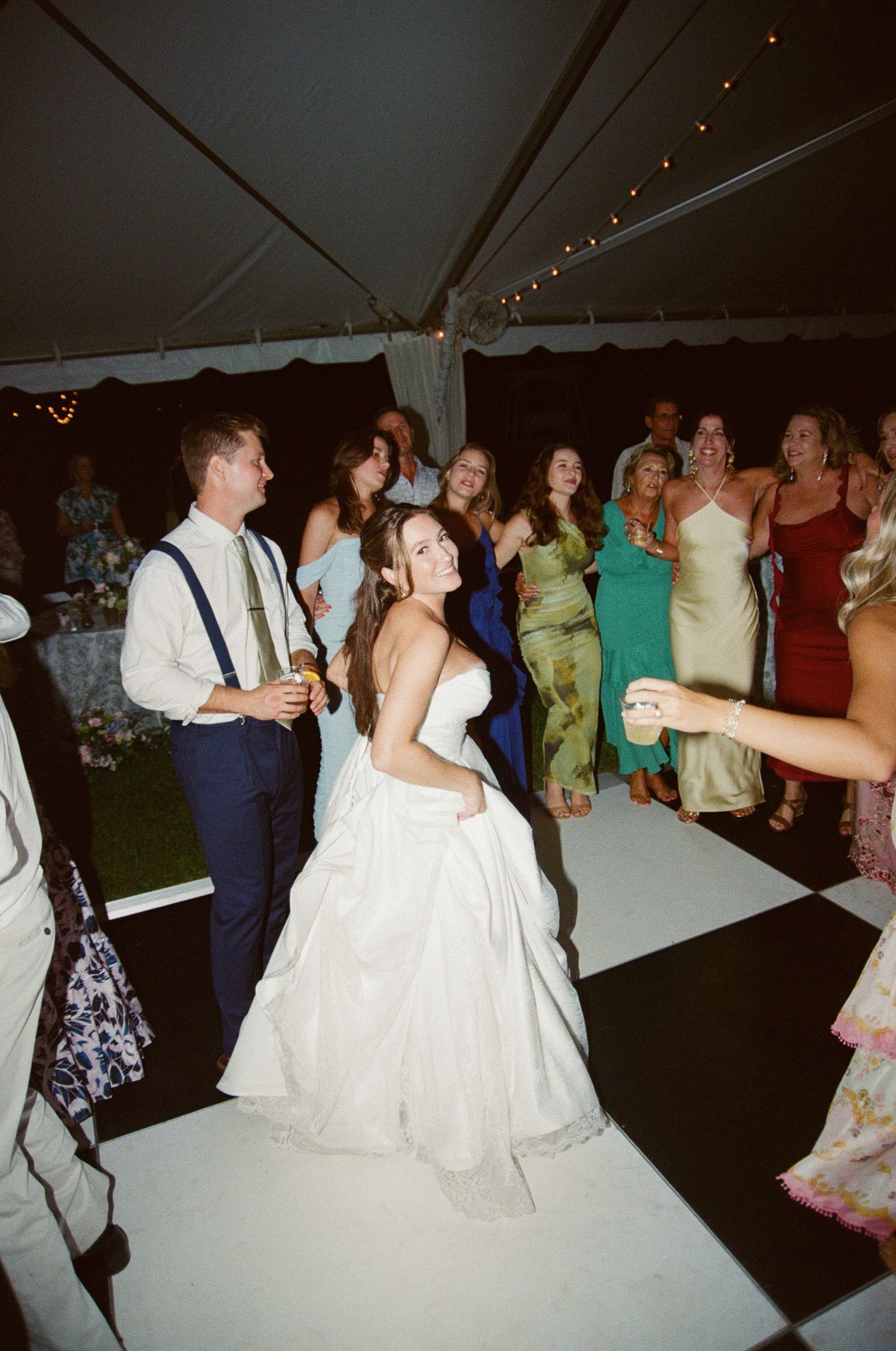 People dancing and celebrating at a wedding reception under a white tent with colorful dresses and formal attire.