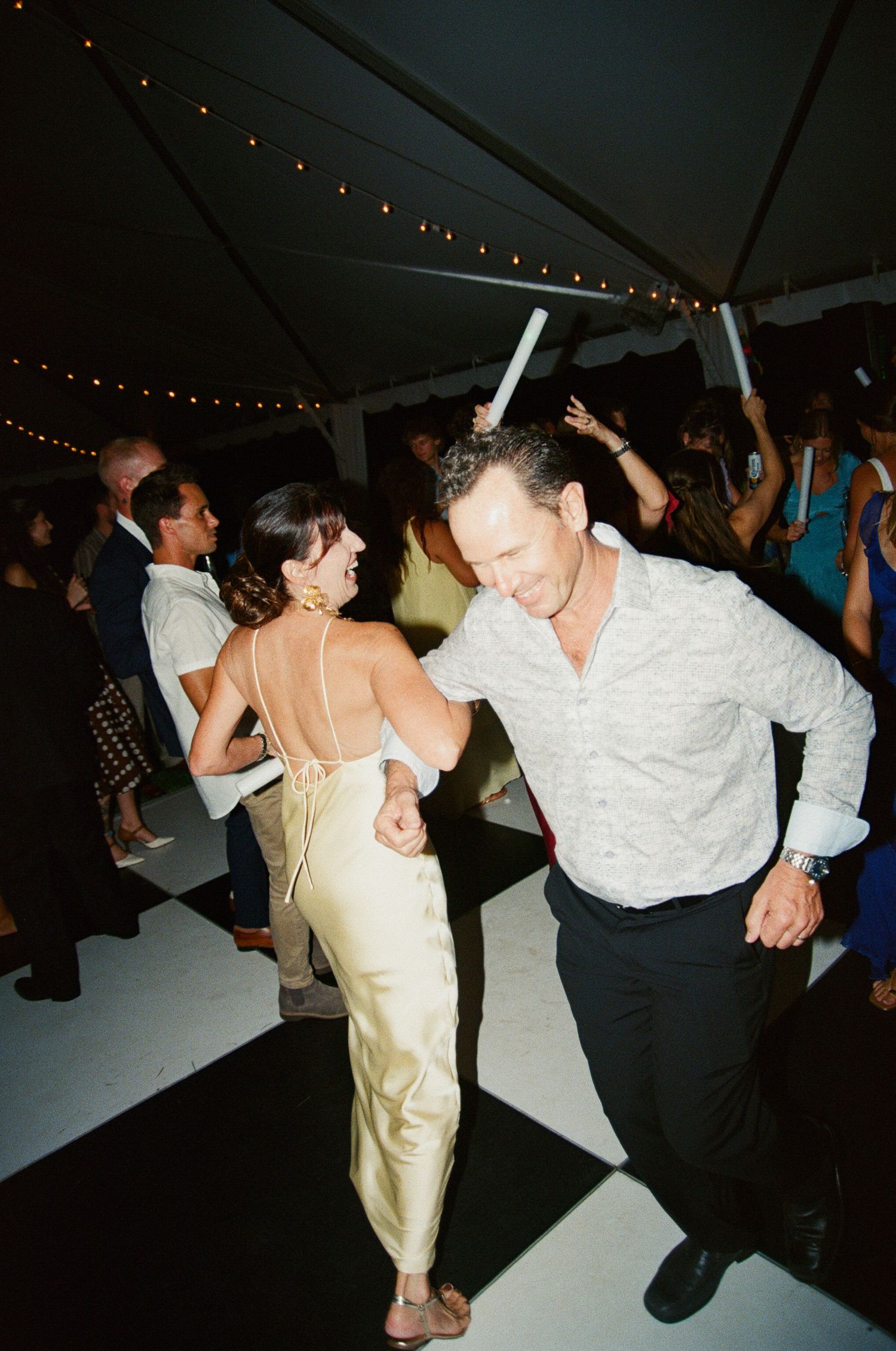 Couples dance together under string lights at an evening reception in a white tent.