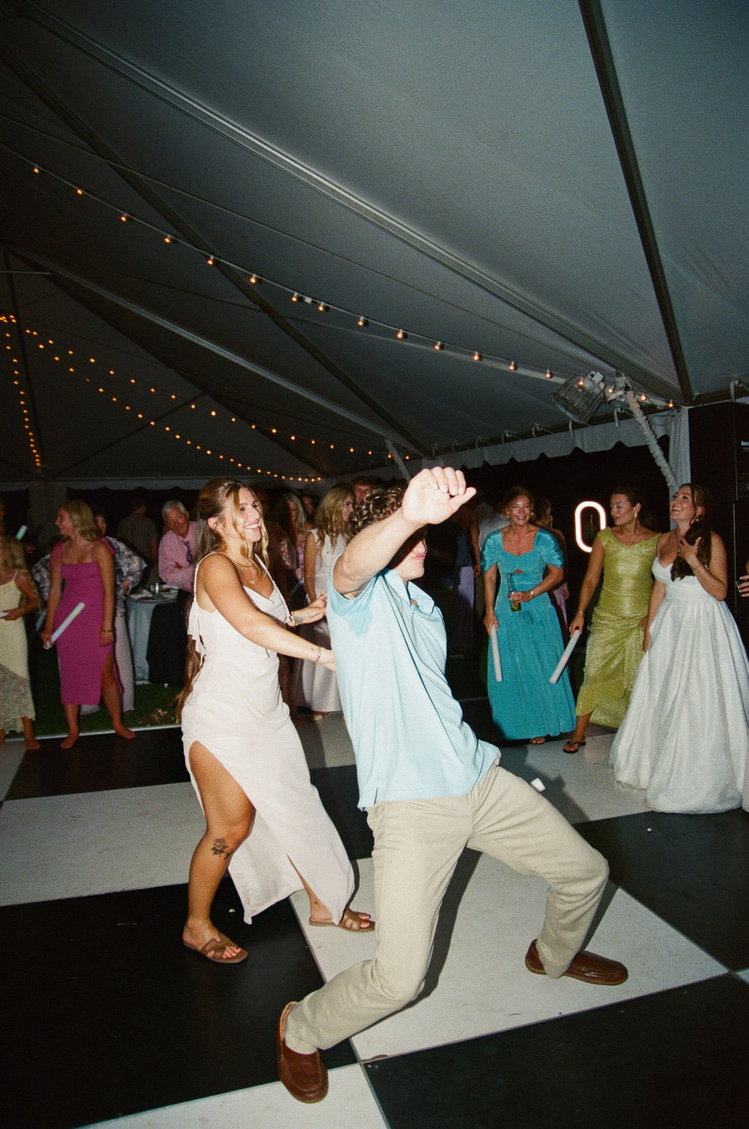 Wedding reception guests dance and celebrate on a dark dance floor under a white tent with string lights overhead.