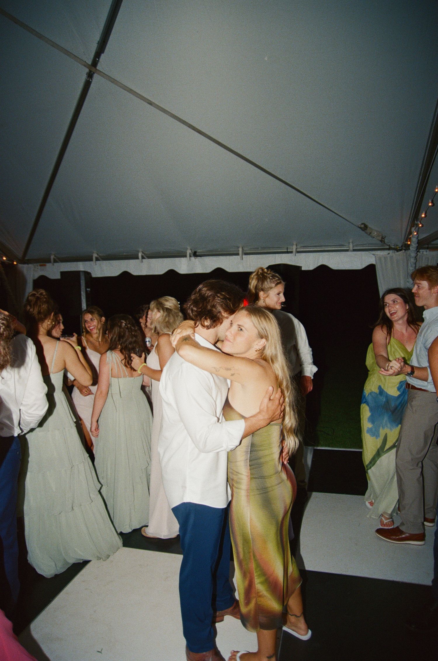 Wedding guests dance together under a white tent during an evening reception celebration.