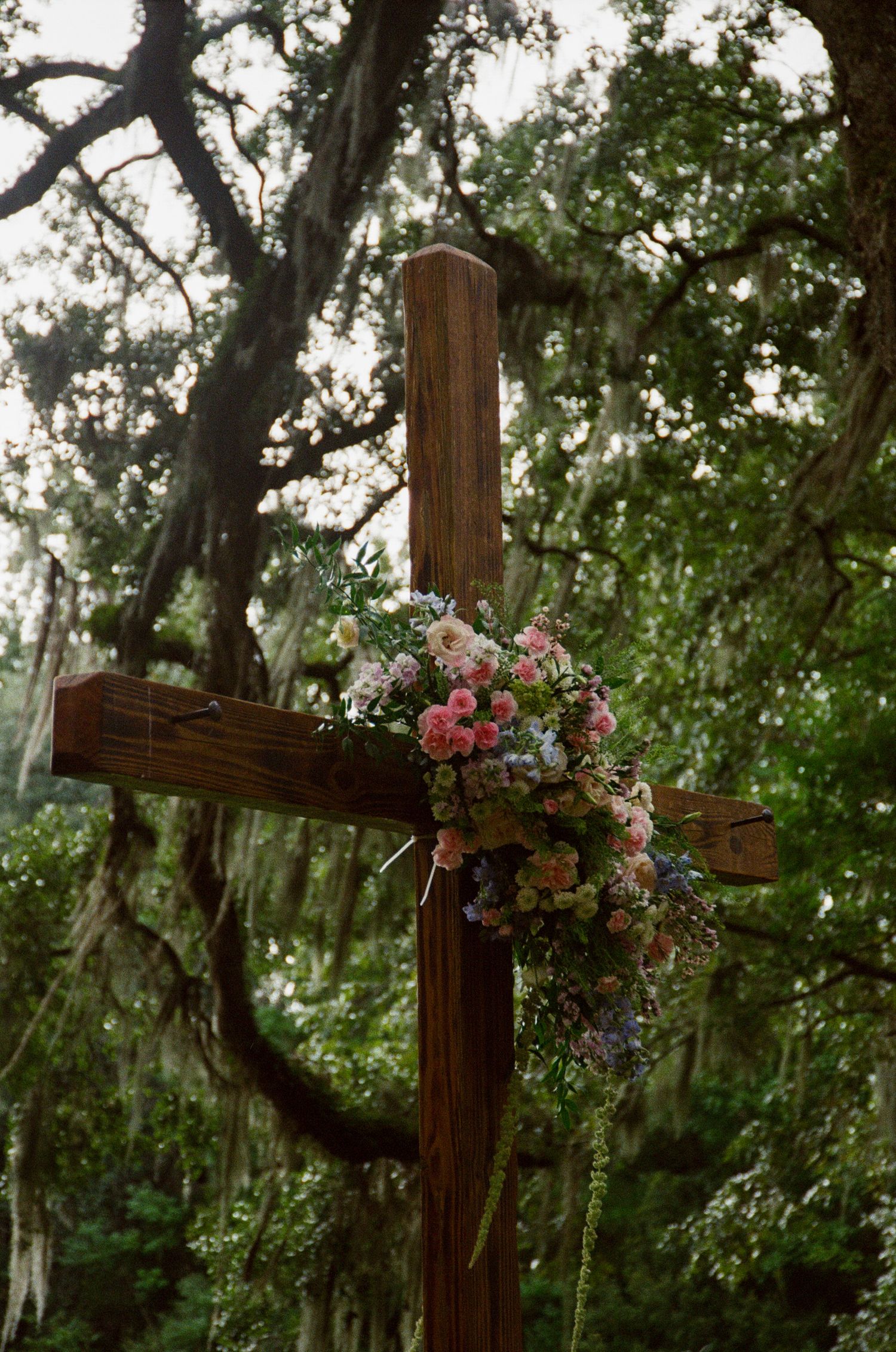 A wooden cross decorated with pink and white flowers stands beneath moss-draped oak trees in a peaceful garden setting.