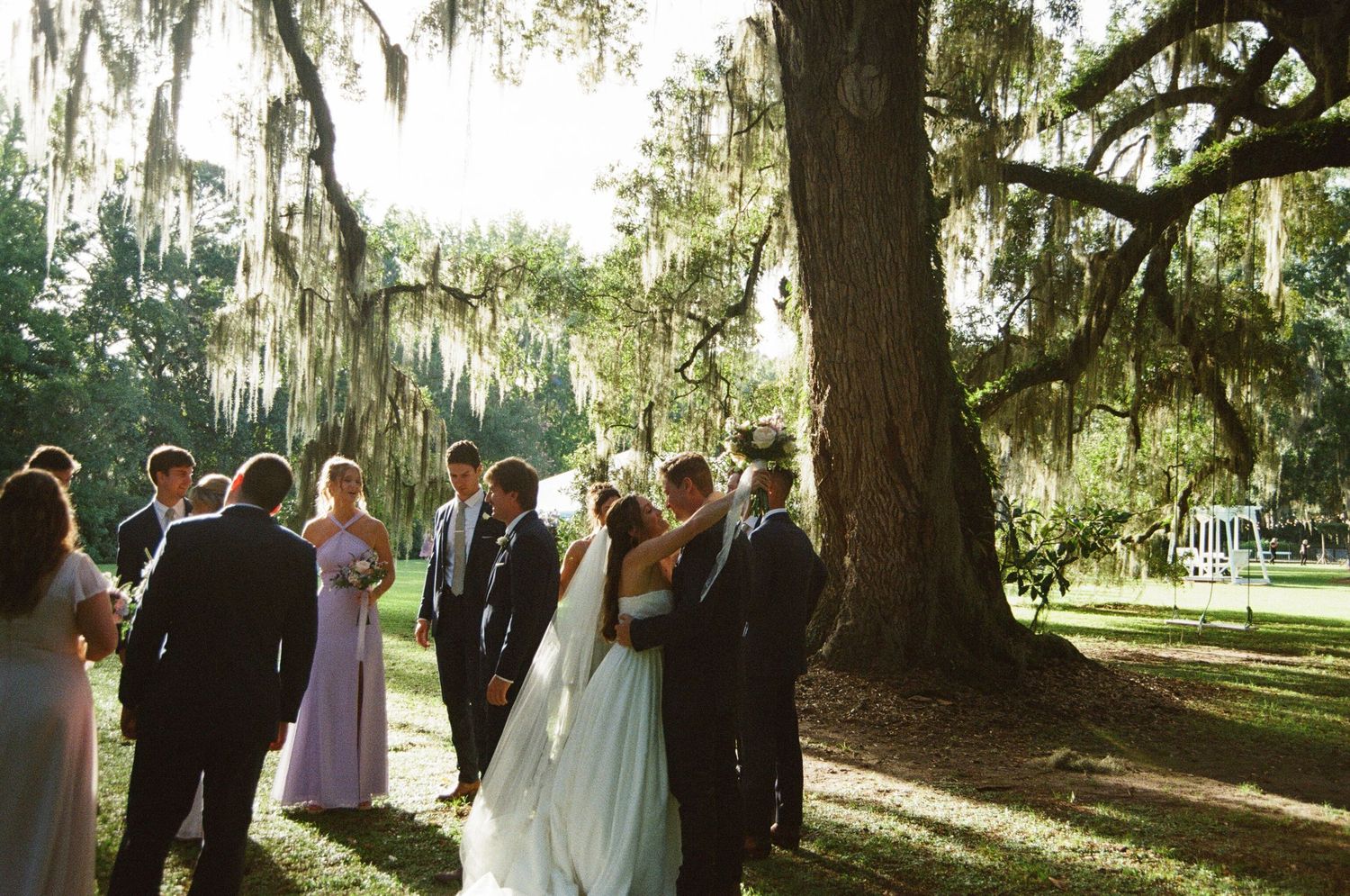 Wedding ceremony takes place under majestic oak trees draped with Spanish moss in a romantic garden setting.
