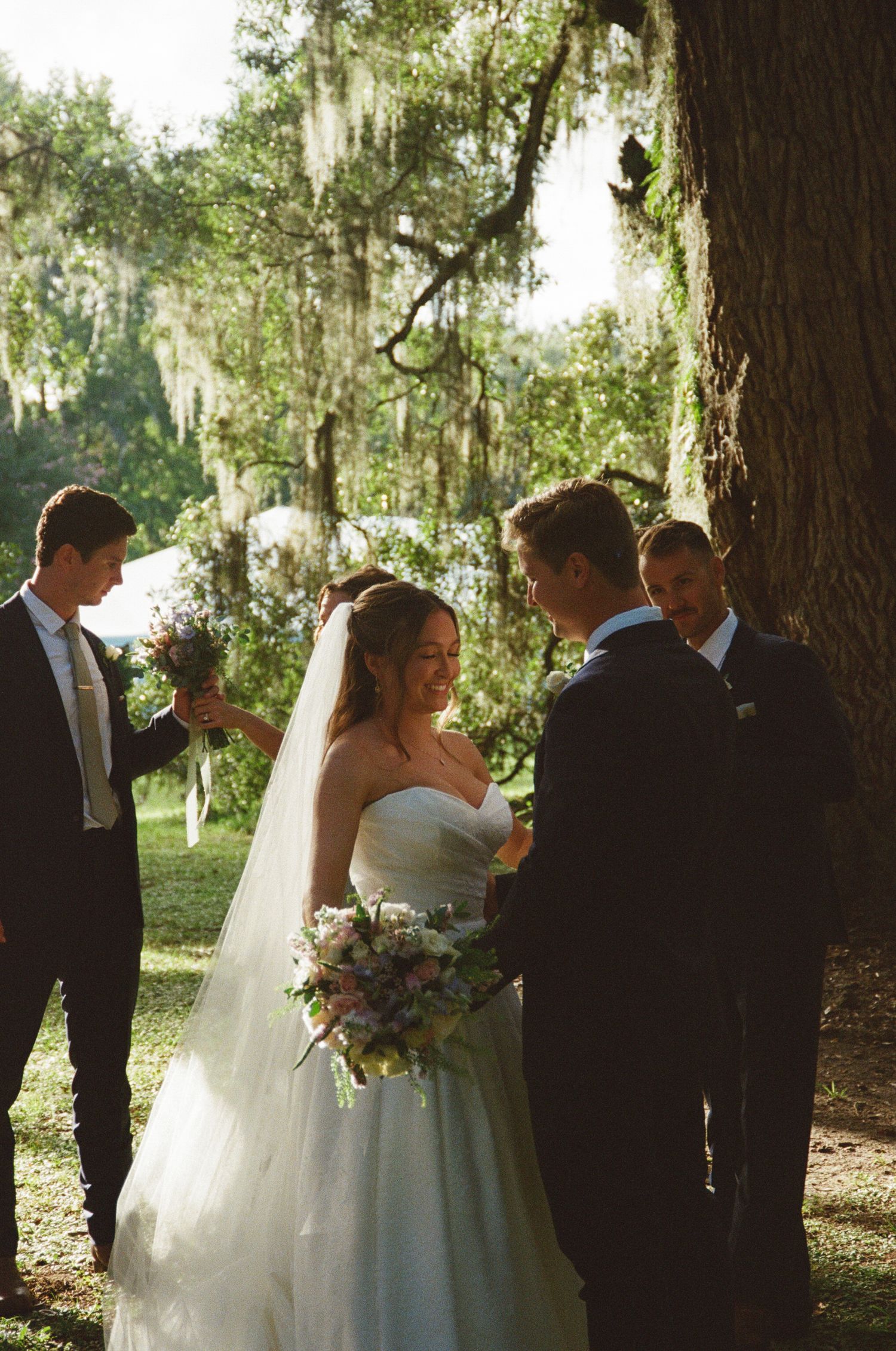 Wedding party gathers under moss-draped oak trees during an outdoor ceremony in natural sunlight.