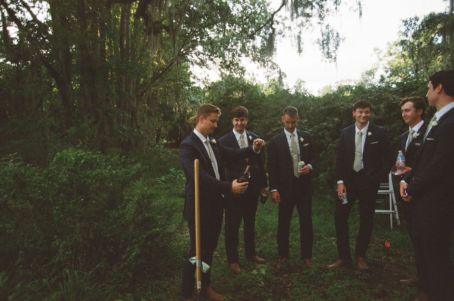 A wedding party stands in a line outdoors under tall trees during a garden ceremony.