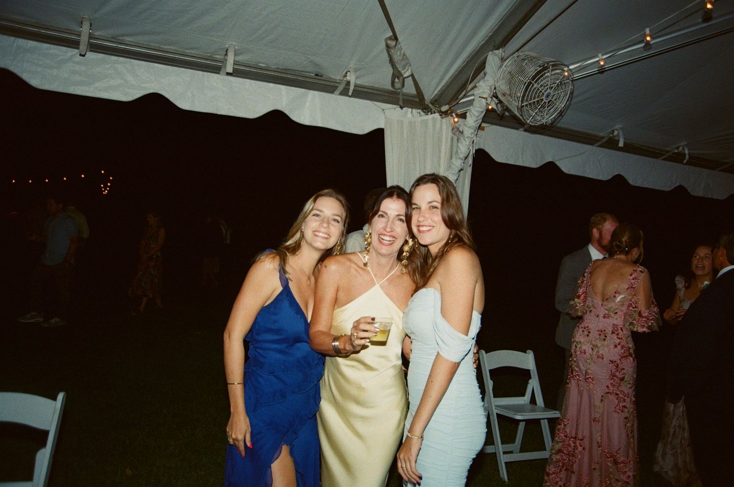 A group of people in formal attire dancing and celebrating at a wedding reception under a white tent.